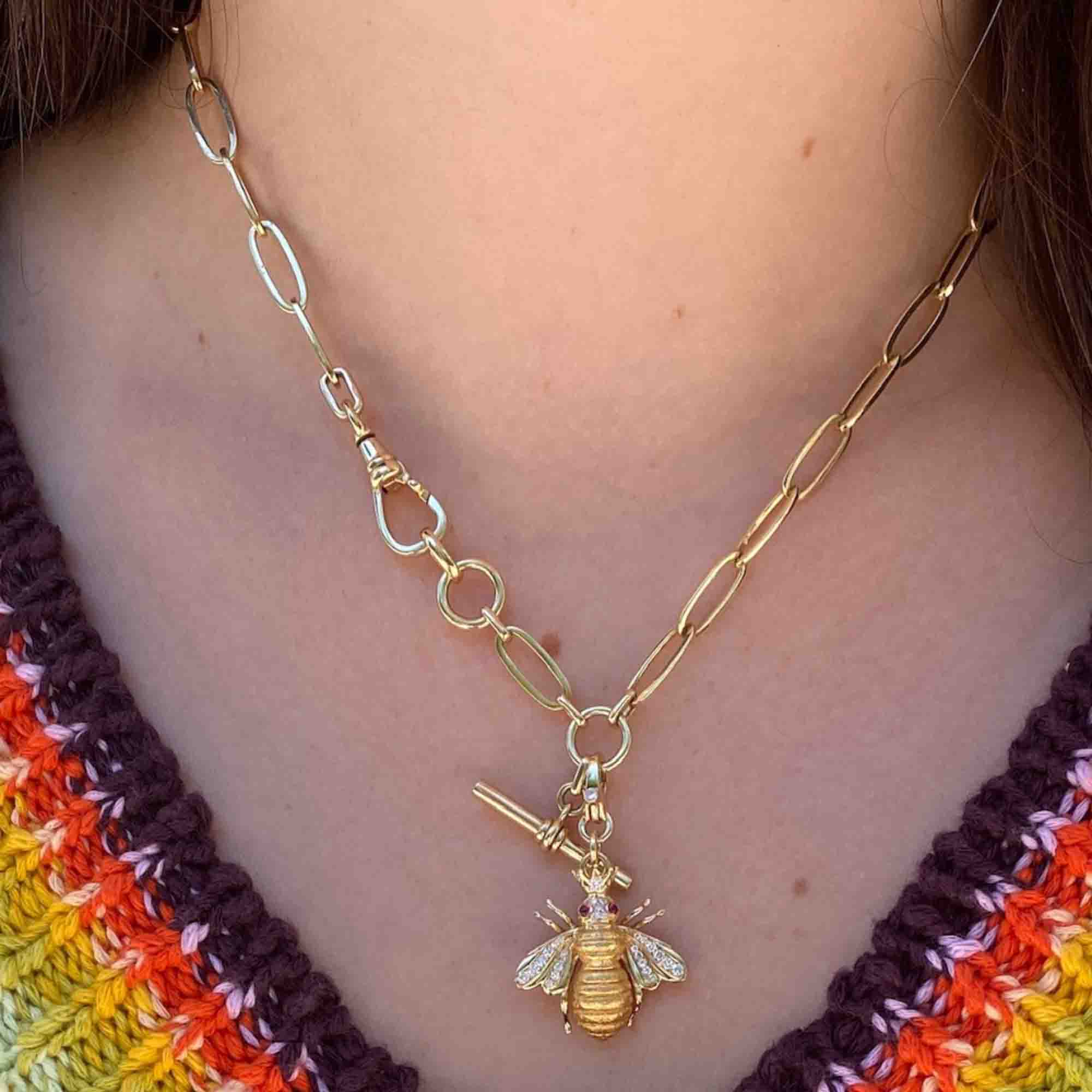 A person wears a Ruby & Diamond Bee Charm on a gold chain necklace with a toggle clasp from Janet Heller Fine Jewelry, complementing their multicolored striped top in shades of purple, yellow, and orange.