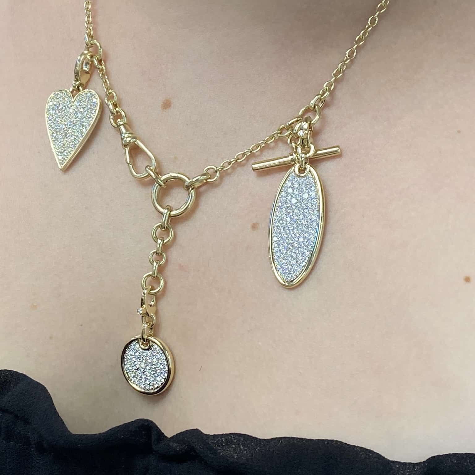 Explore the Berlin Chain by Janet Heller Fine Jewelry, an exquisite gold necklace with heart, circle, and oval charms featuring sparkling textures. This elegant piece has a toggle clasp that beautifully complements black fabric.