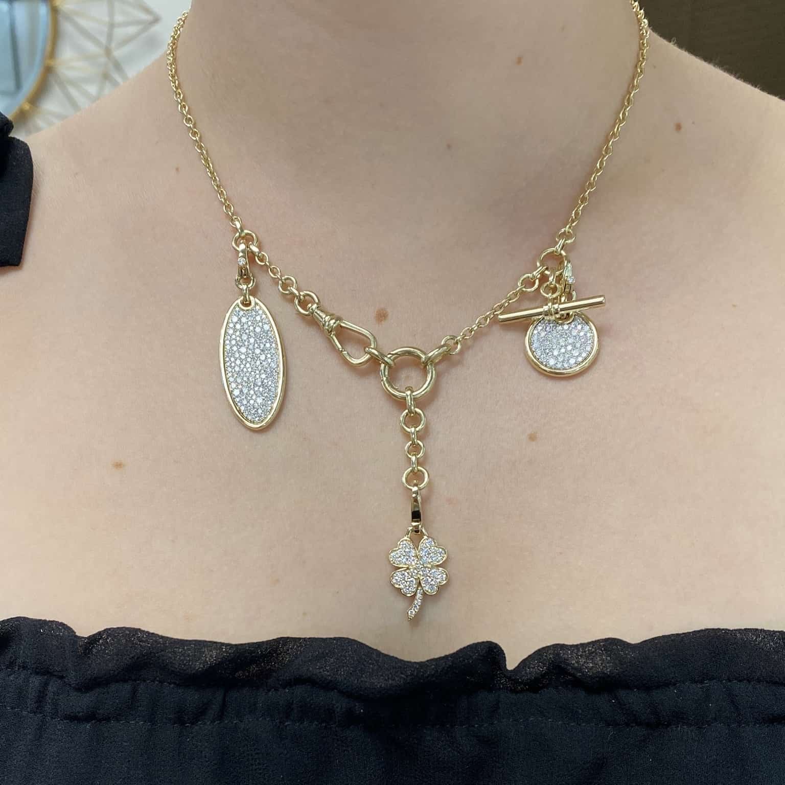 A person wearing a Berlin Chain from Janet Heller Fine Jewelry showcases a clover-shaped pendant with oval and circular charms. The elegant necklace, featuring a toggle clasp, perfectly complements the black top with ruffled edges.