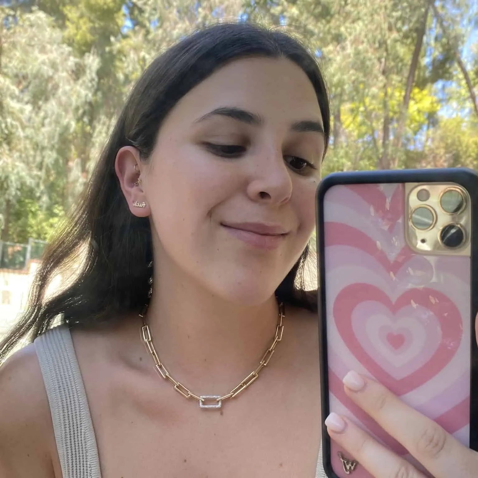 A person with long dark hair takes a selfie outdoors, wearing exquisite Janet Heller Fine Jewelry—a Rectangular Diamond Link Necklace and earrings. Their phone case has a pink heart design, with trees and sunlight creating a serene backdrop.