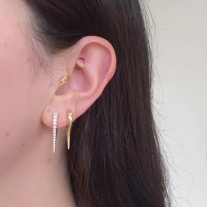 A close-up of a persons ear showcases exquisite jewelry: a custom gold lightning bolt stud at the top, a gold hoop in the cartilage, Diamond Dagger Earrings with sparkling stones forming a dangle earring, and a gold curved bar below.