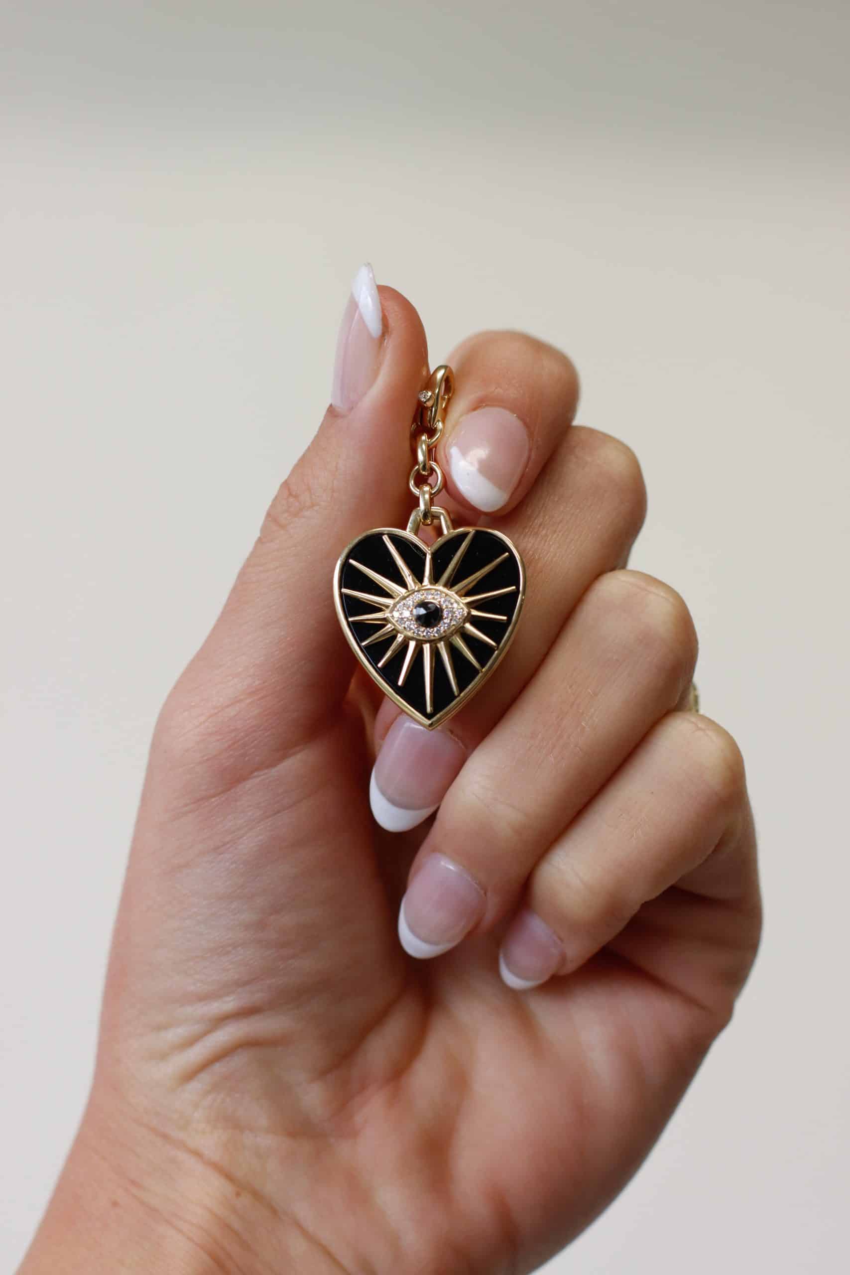 The Black Agate Heart Burst Charm from Janet Heller Fine Jewelry is crafted in 14KT gold. The charm is set in black enamel with gold rays. The center of the pendant features an evil eye with a center black diamond that is surrounded by white diamonds. The diamond clasp clips easily onto various Janet Heller Fine Jewelry chains and necklaces.