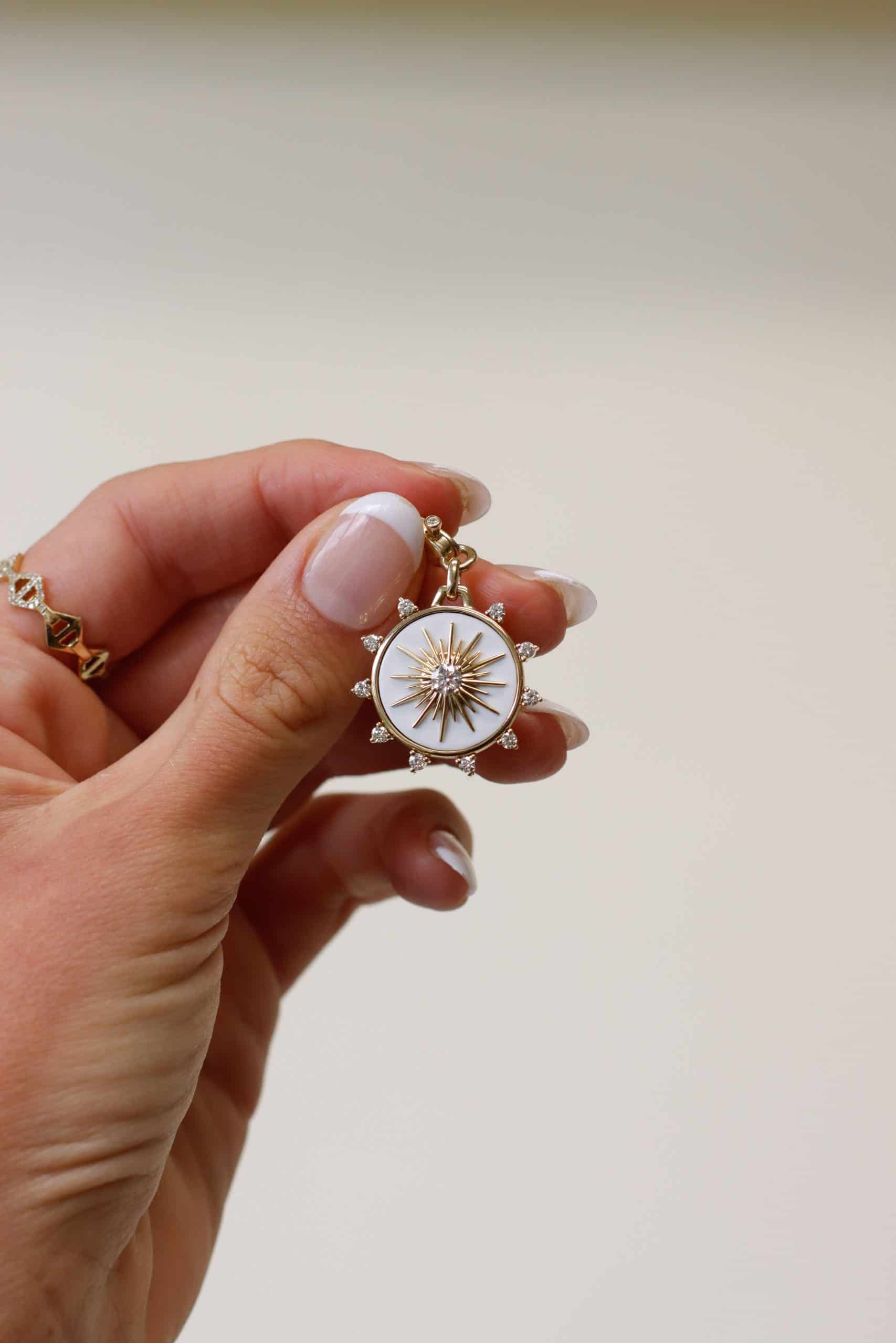 A person with manicured nails holds the Diamond & Cacholong Bentley Charm from Janet Heller Fine Jewelry, featuring a white center and radiating star design. Surrounding the star are small sparkling stones set in a metallic frame, offering endless customizations typical of exquisite jewelry.