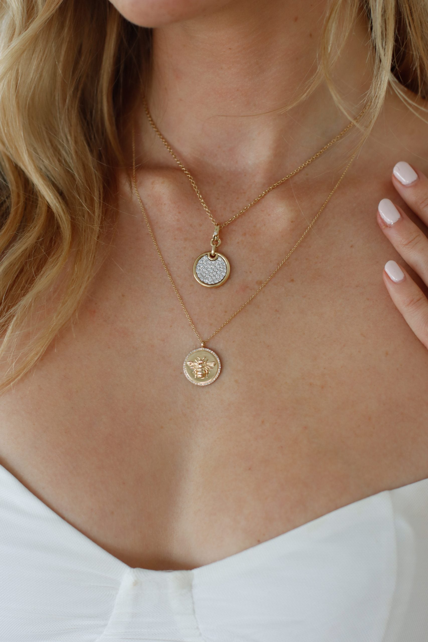 A woman in Janet Heller Fine Jewelry wears a gold necklace featuring a Diamond Bee Pendant and an intricately designed one. Her white off-shoulder top and light pink nails complete the look.