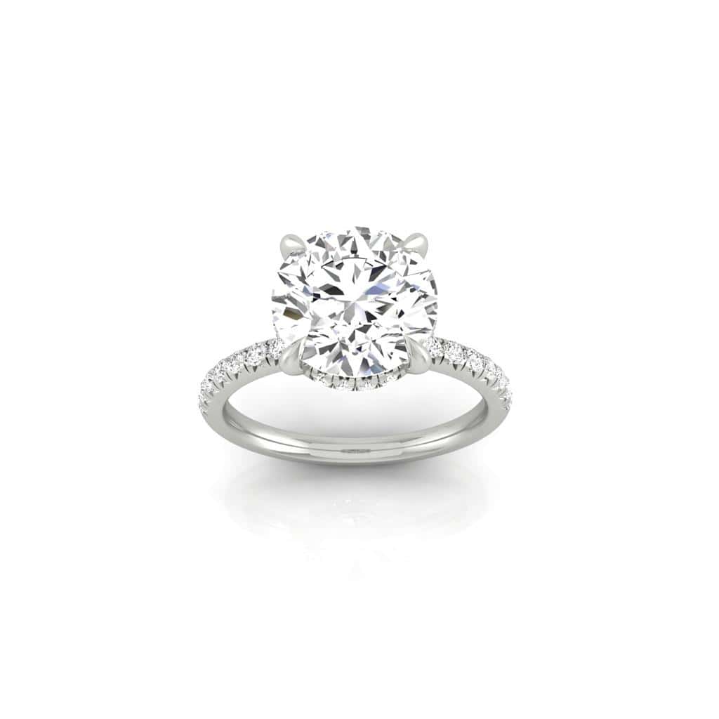 The Amélie + Hidden Halo is a silver engagement ring featuring a large round clear diamond with an elegant hidden halo design. The French Pavé set band sparkles with smaller white stones, shining brilliantly on a white background.