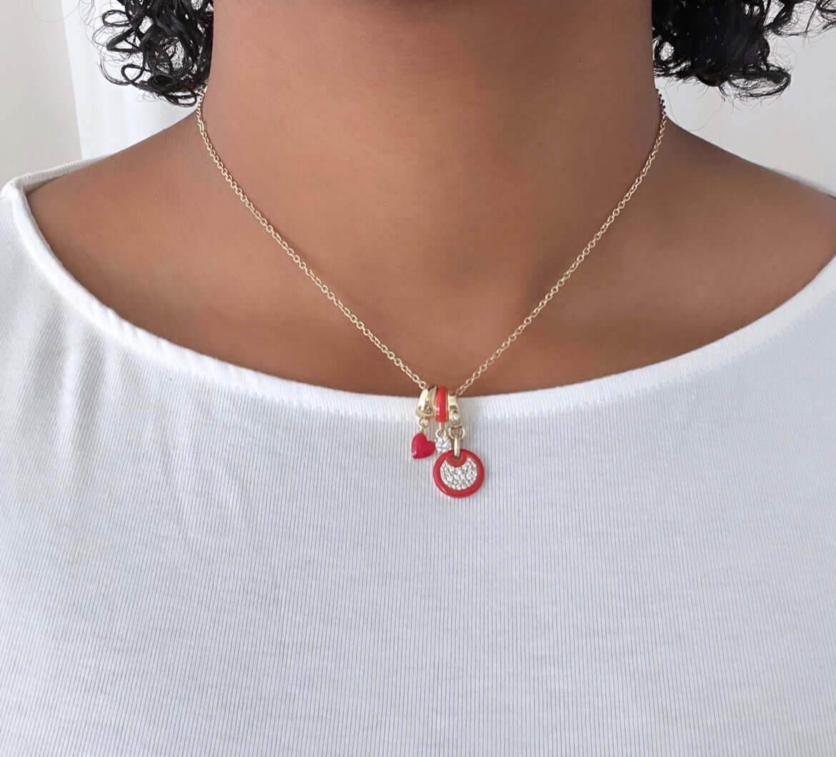 Wearing a Janet Heller Fine Jewelry gold necklace with customized charms—a small red heart, key, and inscribed “LOVE”—perfectly complements their white top. Featured: Red Enamel Mini Round Diamond Charm.