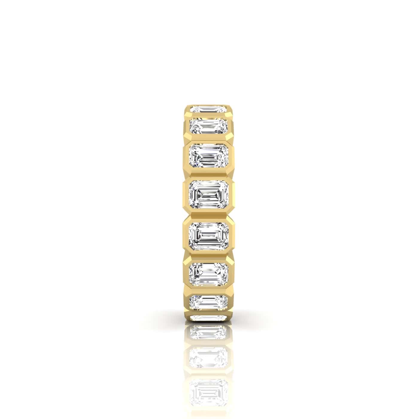 The Bezel Set Emerald Cut Eternity Ring features an 18KT gold band adorned with symmetrical rectangular emerald-cut diamonds of VS clarity and F color, set in a continuous line for a beautifully reflective and elegant design.