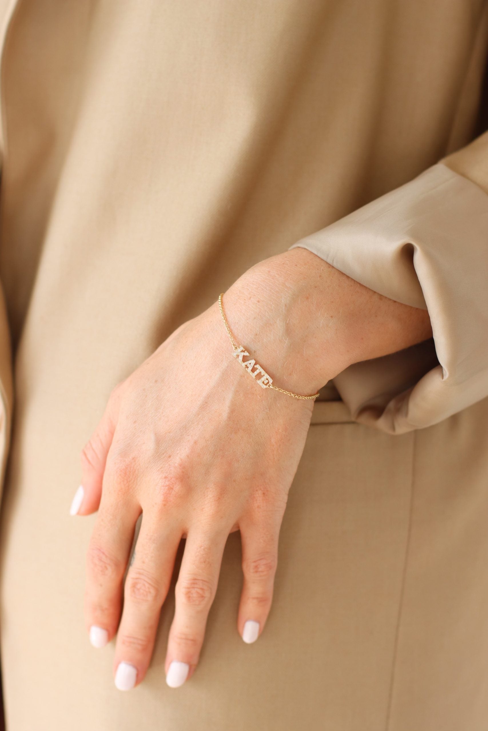 A person dressed in beige displays their left hand, adorned with the elegant Diamond Block Name Bracelet engraved with KATE, embodying the charm of personalized jewelry. Their neatly manicured nails are coated in light pink polish.