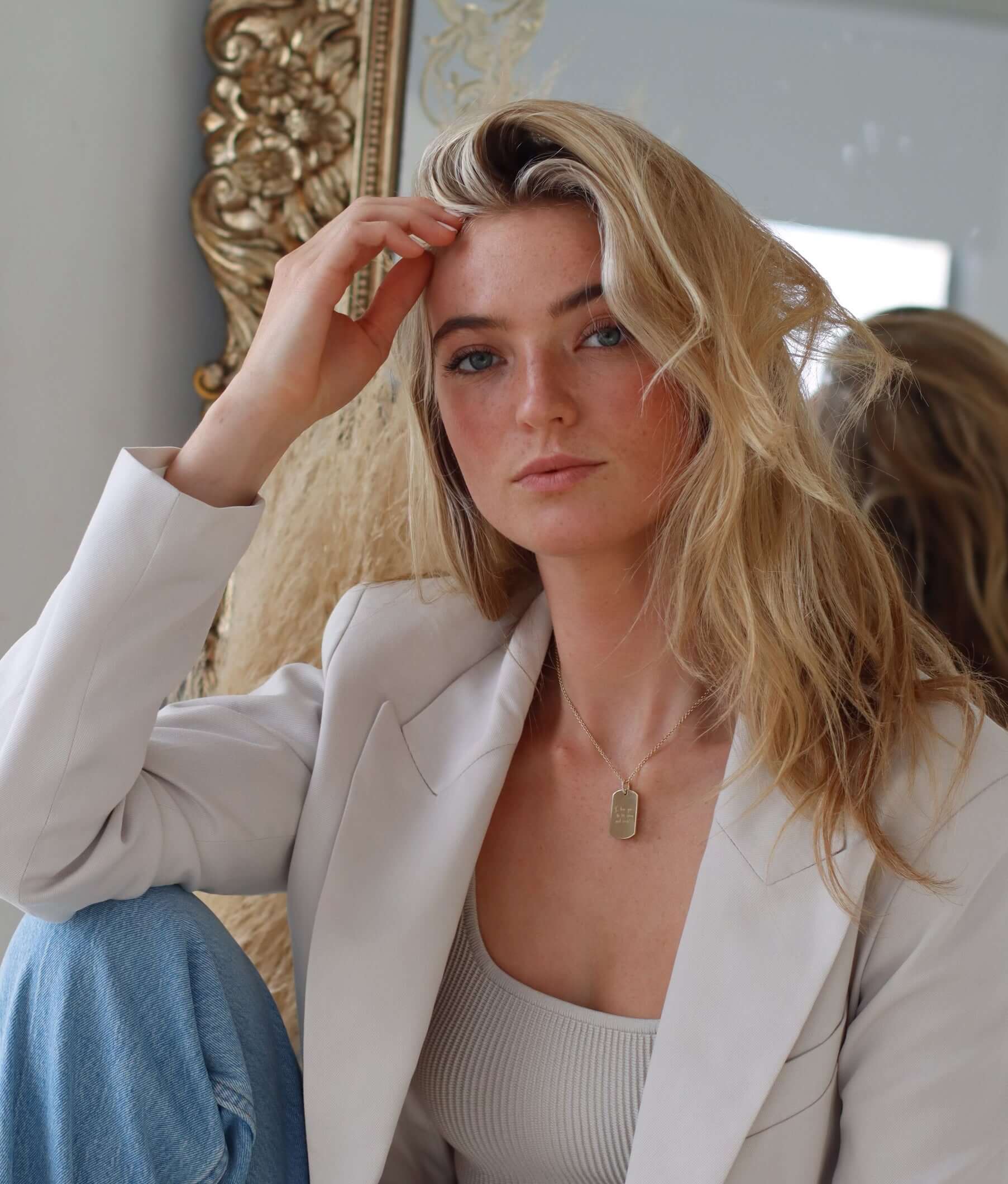 A person with long blonde hair sits casually, wearing a white blazer and ribbed top, accessorized with a Large Dog Tag Engraveable Pendant Necklace. One hand rests on their head as they gaze at the camera. A mirror with an ornate frame adds warmth and character to the scene.