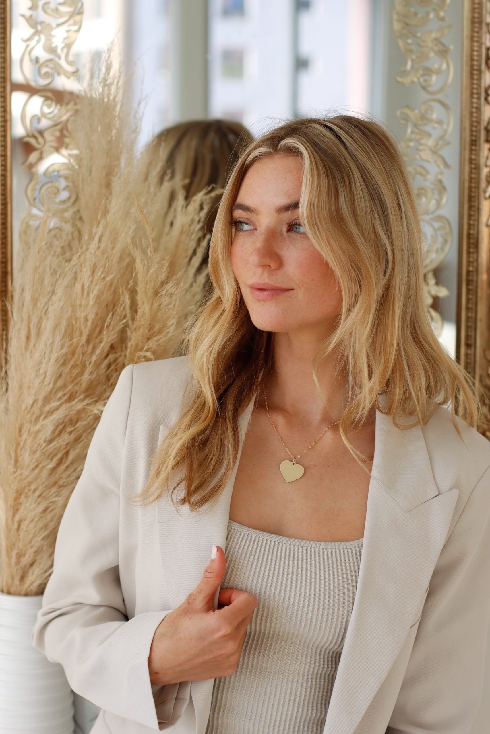 A woman with long blonde hair and a gentle expression stands in front of a mirror, wearing a cream blazer and a Large Heart Engraveable Pendant Necklace. Beside her is pampas grass in a white vase as she gazes to the right.