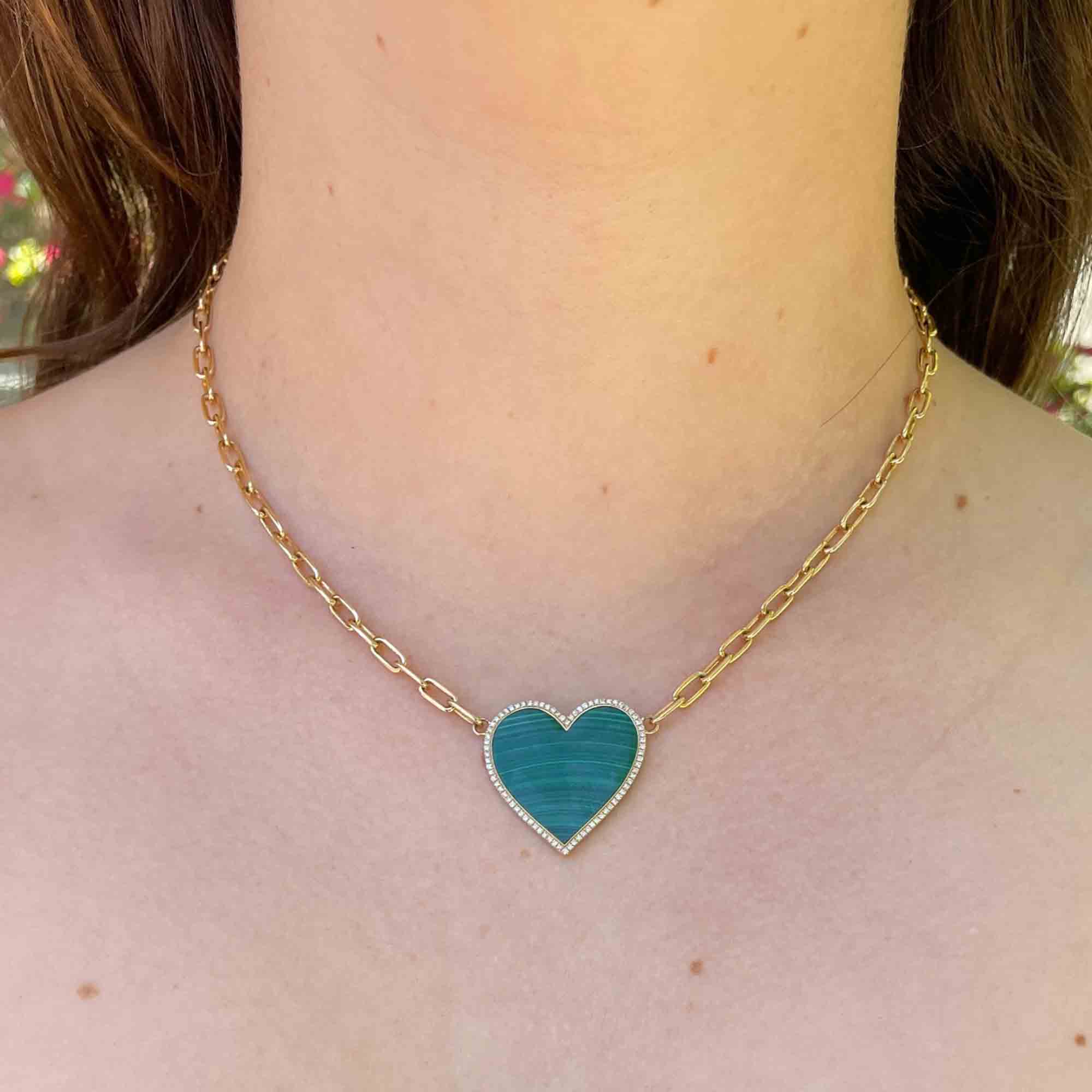 A woman wears a Paperclip Link Malachite Heart Necklace by janethellerfinejewelry, featuring a shiny golden chain and a blue heart-shaped pendant. It sits elegantly centered on her neckline against a softly blurred background, enhancing its fine jewelry allure.