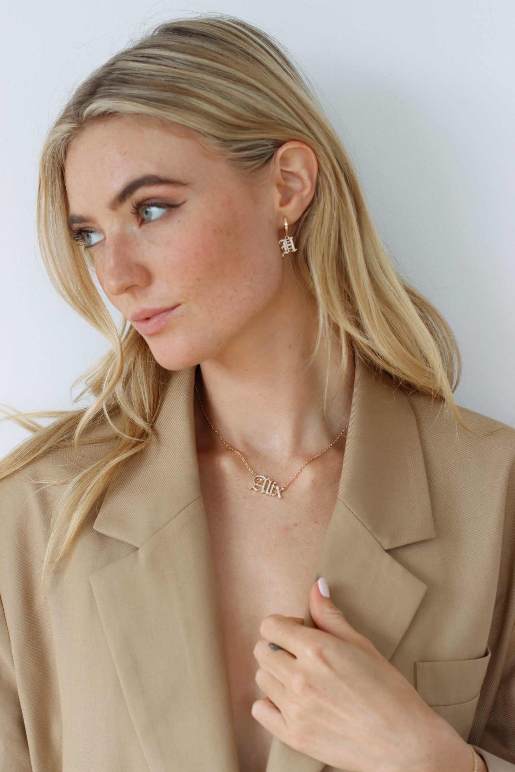 A woman with long blonde hair and subtle makeup, highlighting her eyeliner, gazes to the side while wearing a beige blazer. She accessorizes with unique jewelry, including the gold Small Old English Initial Huggies. The background is plain white.
