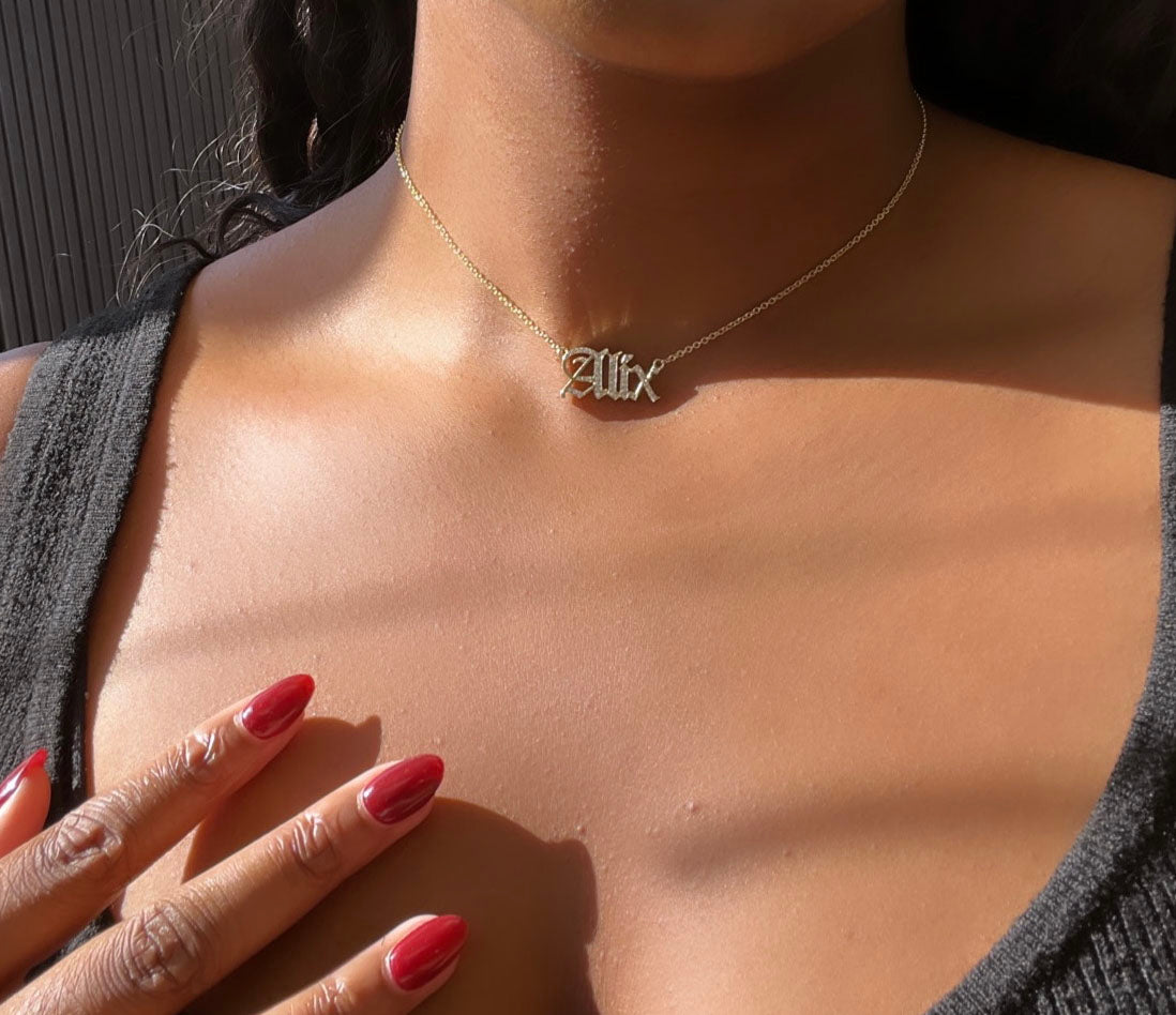 A person in a dark top, with manicured red nails gently touching their collarbone, wears the Diamond Old English Name Necklace, featuring a pendant with script lettering spelling Alix, perfectly capturing the allure of personalized jewelry.