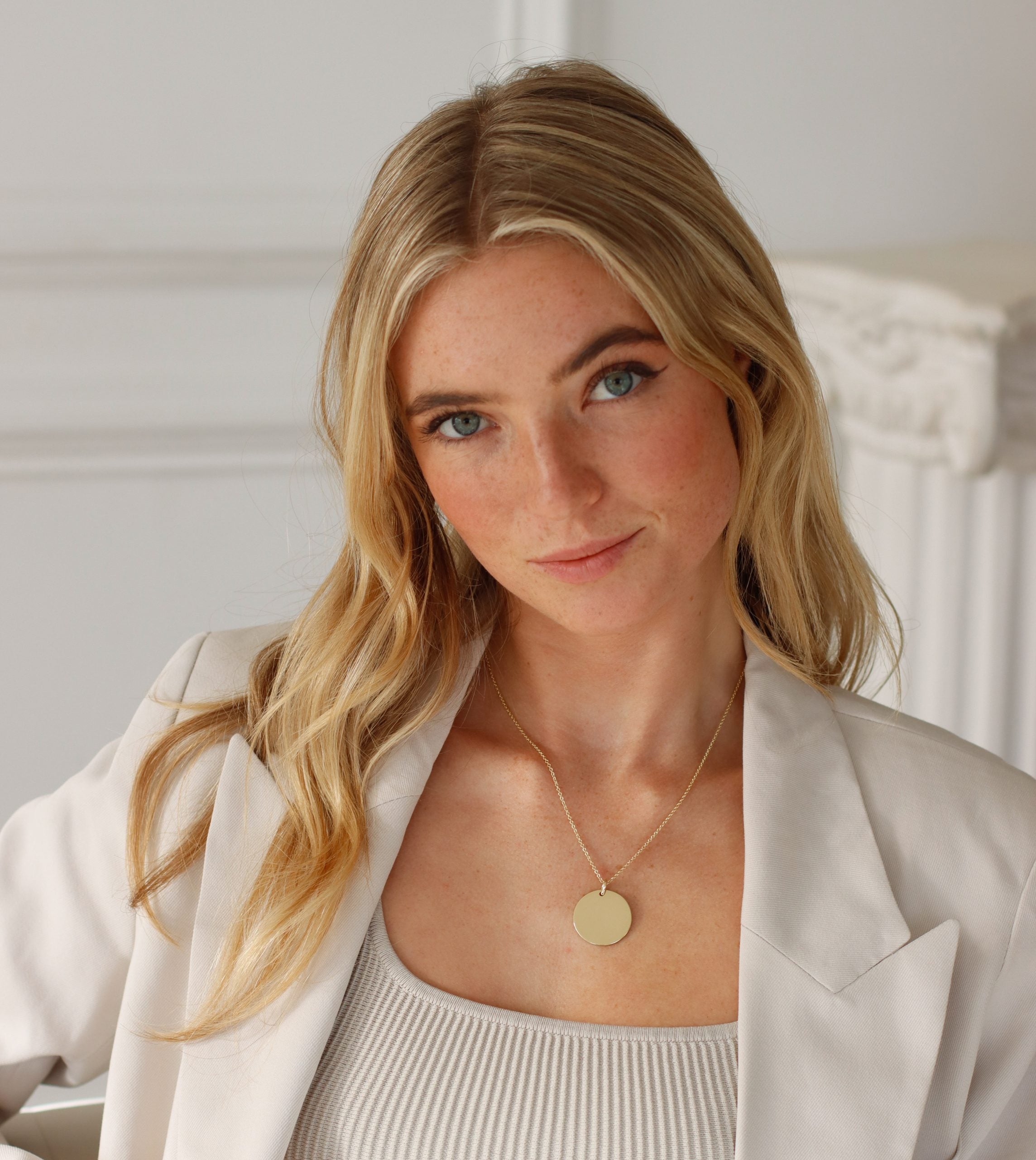 A person with long blonde hair and blue eyes, dressed in a beige blazer over a white top, wears a Large Round Engraveable Pendant Necklace. They look at the camera with a slight smile against a softly lit interior background.