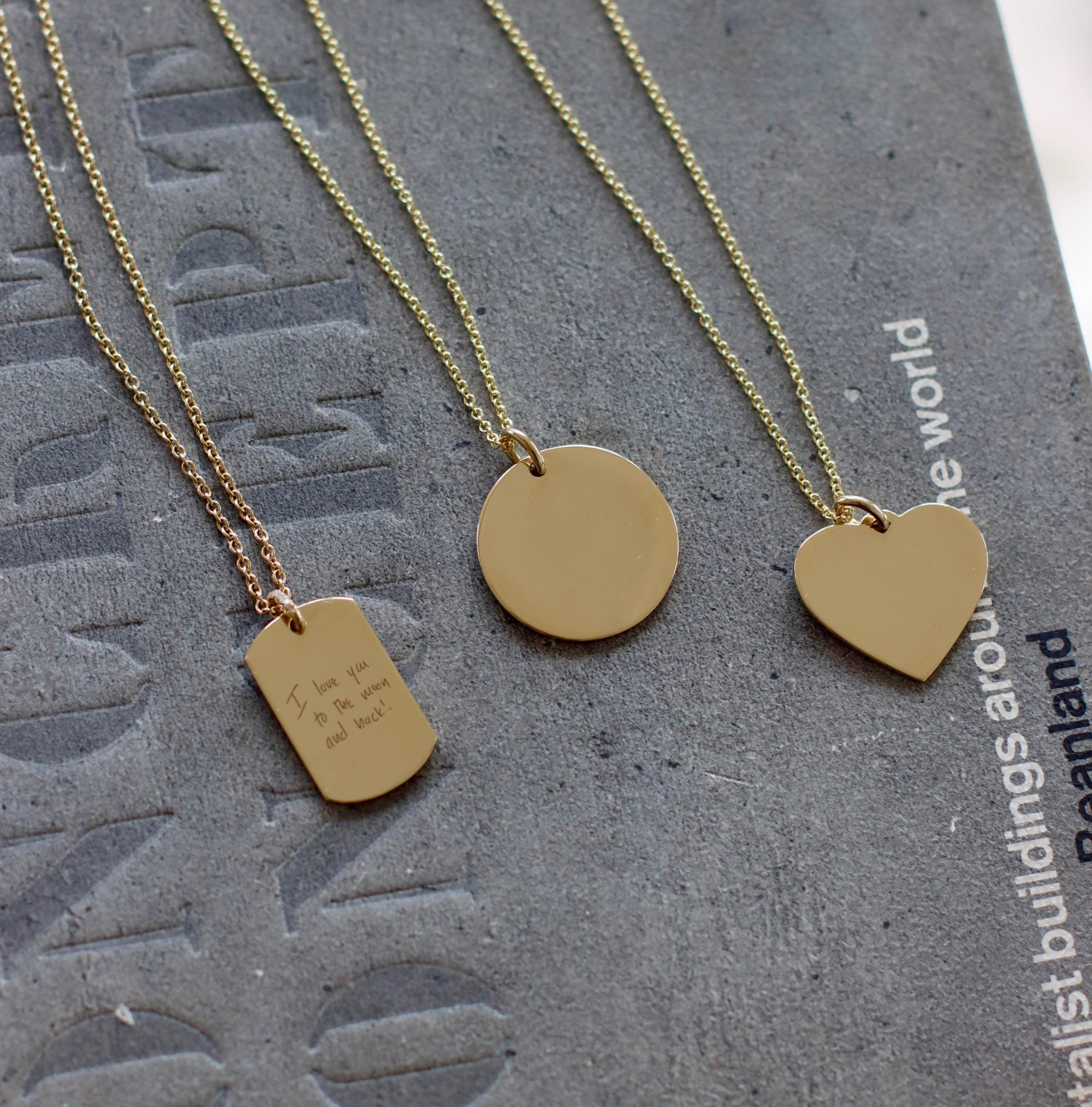 Three gold necklaces lie on a textured surface: a vertical rectangle with custom engraving, a circular disc, and the Large Heart Engraveable Pendant Necklace. Partially visible text in the background elevates their charm as personalized keepsakes.