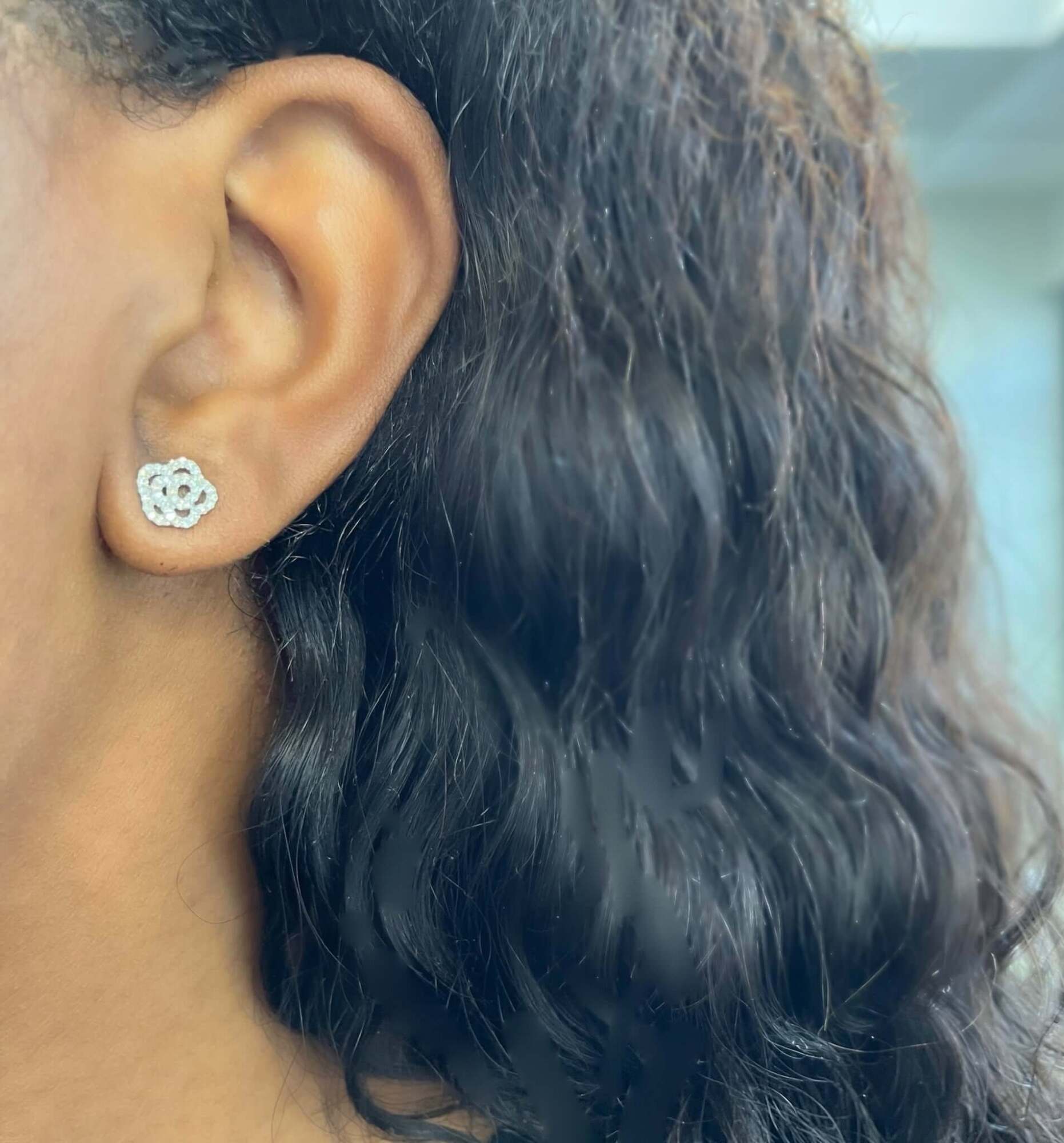A close-up of a person’s ear with long, wavy dark hair showcases the elegance of fine jewelry with the Diamond Rose Studs—sparkling, flower-shaped earrings crafted from small stones.