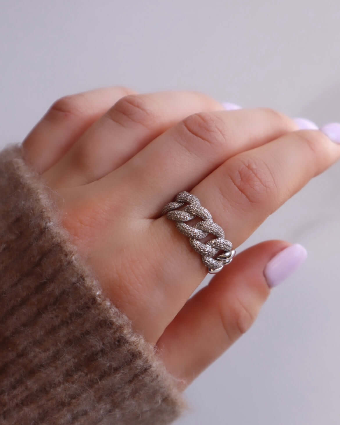 A hand with light purple nail polish showcases the Diamond Hunter Ring—a chunky silver chain-link design worn on the middle finger for a bold, standout look that merges fine jewelry with everyday style. Paired with a soft brown knitted sweater, this eye-catching accessory is available for preorder, so you can choose your perfect ring size for the ideal fit.