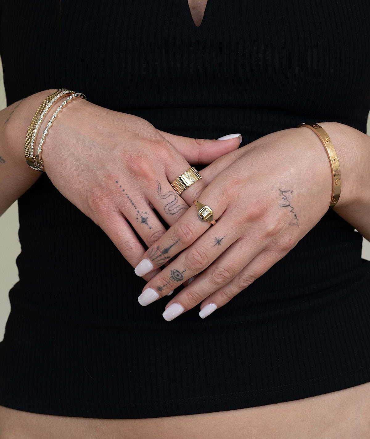 A person with light skin and white manicured nails wears the Geo Cut Baguette Diamond Signet Ring, displaying hand and finger tattoos—lines, dots, a star, and an eye—against a ribbed black top.