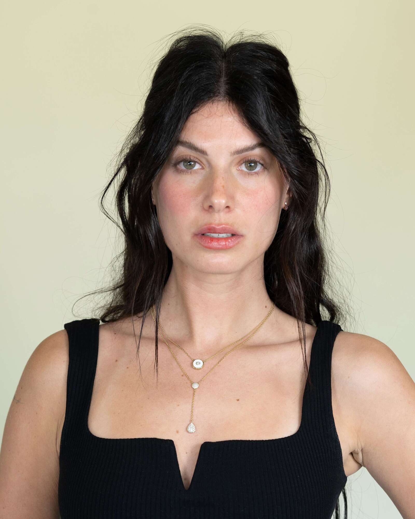 A woman with long dark wavy hair wears a black sleeveless top and the Diamond Teardrop & Round Lariat Necklace from Janet Heller Fine Jewelry, standing against a plain light background and looking directly at the camera.