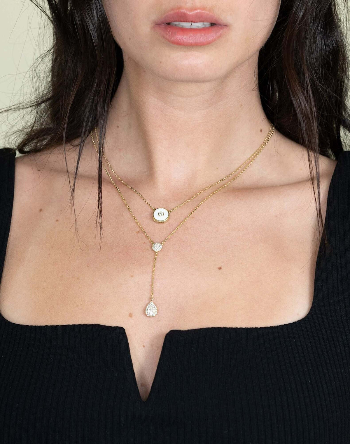 A woman in a black V-neck top showcases the Diamond Teardrop & Round Lariat Necklace by Janet Heller Fine Jewelry, featuring layered gold chains with a round pendant and two teardrop pendants, set against a neutral background.