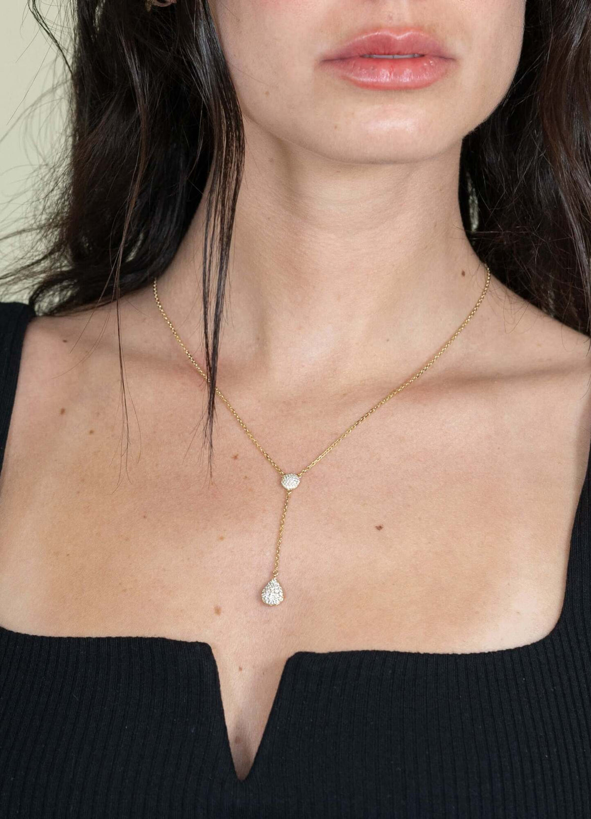 A woman in a black V-neck top models the Janet Heller Fine Jewelry Diamond Teardrop & Round Lariat Necklace, featuring two diamond-studded pendants at different lengths. Her slightly parted lips highlight this elegant custom piece.