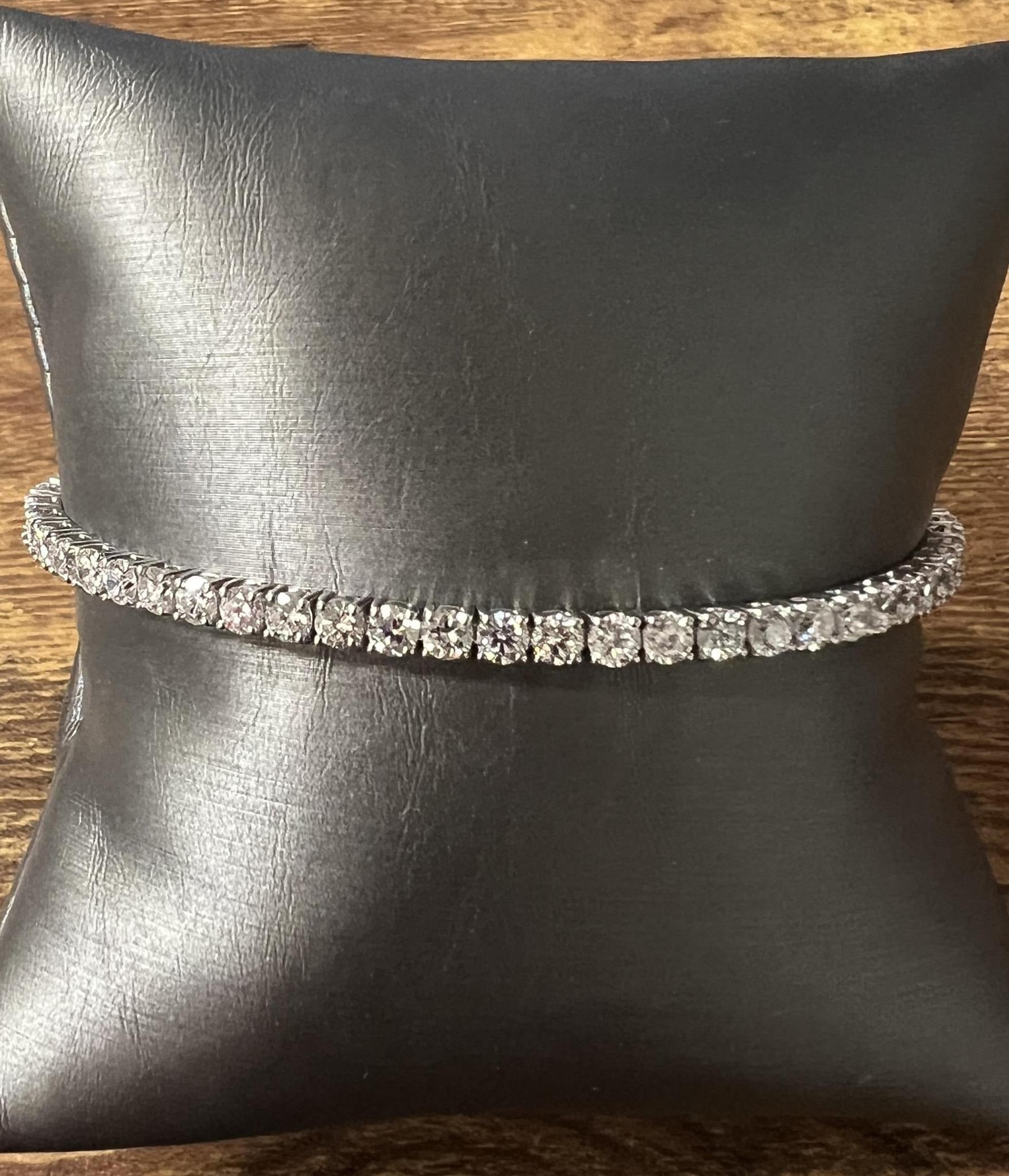 The 14KW 6ctw Lab Diamond Tennis Bracelet (7") with round stones is displayed on a black leather cushion atop a wooden surface.