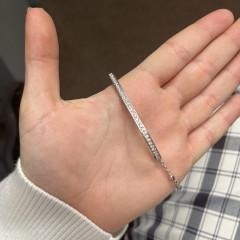 A hand holding the 14KW Curved Diamond Bar Bolo Bracelet, a thin silver bracelet adorned with small diamonds, against a background featuring part of a plaid-patterned garment.