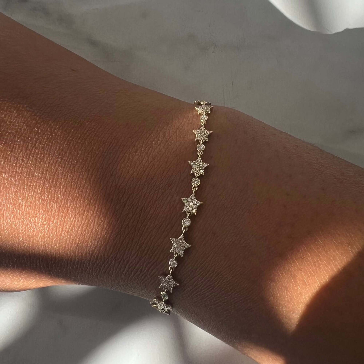 The Repeating Diamond Stars Bracelet from Janet Heller Fine Jewelry, a delicate gold piece adorned with small star-shaped charms, is elegantly worn on the wrist and illuminated by soft natural light, casting gentle shadows on the skin and background. For jewelry inquiries or to explore custom options, please contact us.