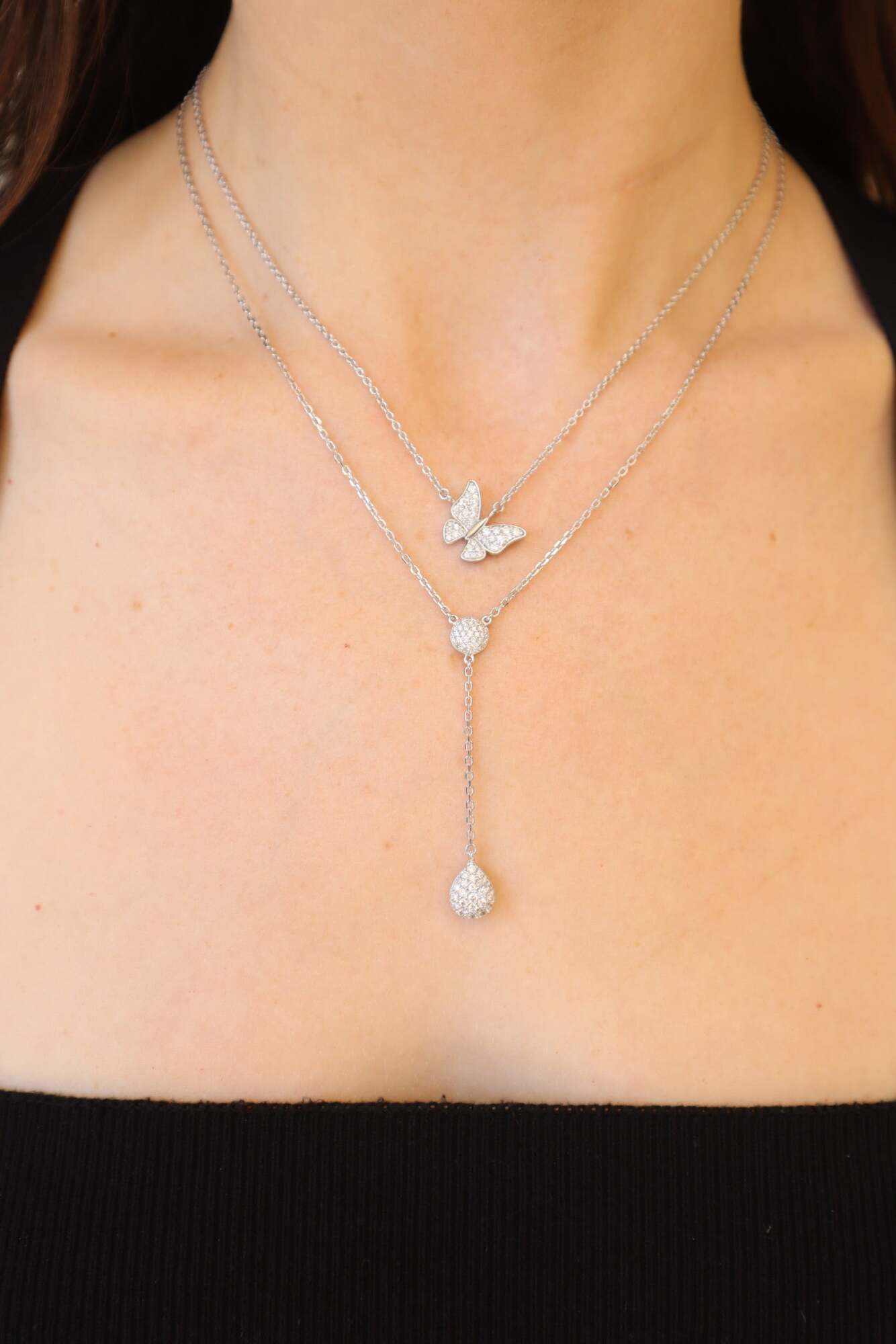 A woman wears Janet Heller Fine Jewelry's Diamond Teardrop & Round Lariat Necklace layered with a butterfly pendant, paired with a black strapless top. For jewelry inquiries, contact us.