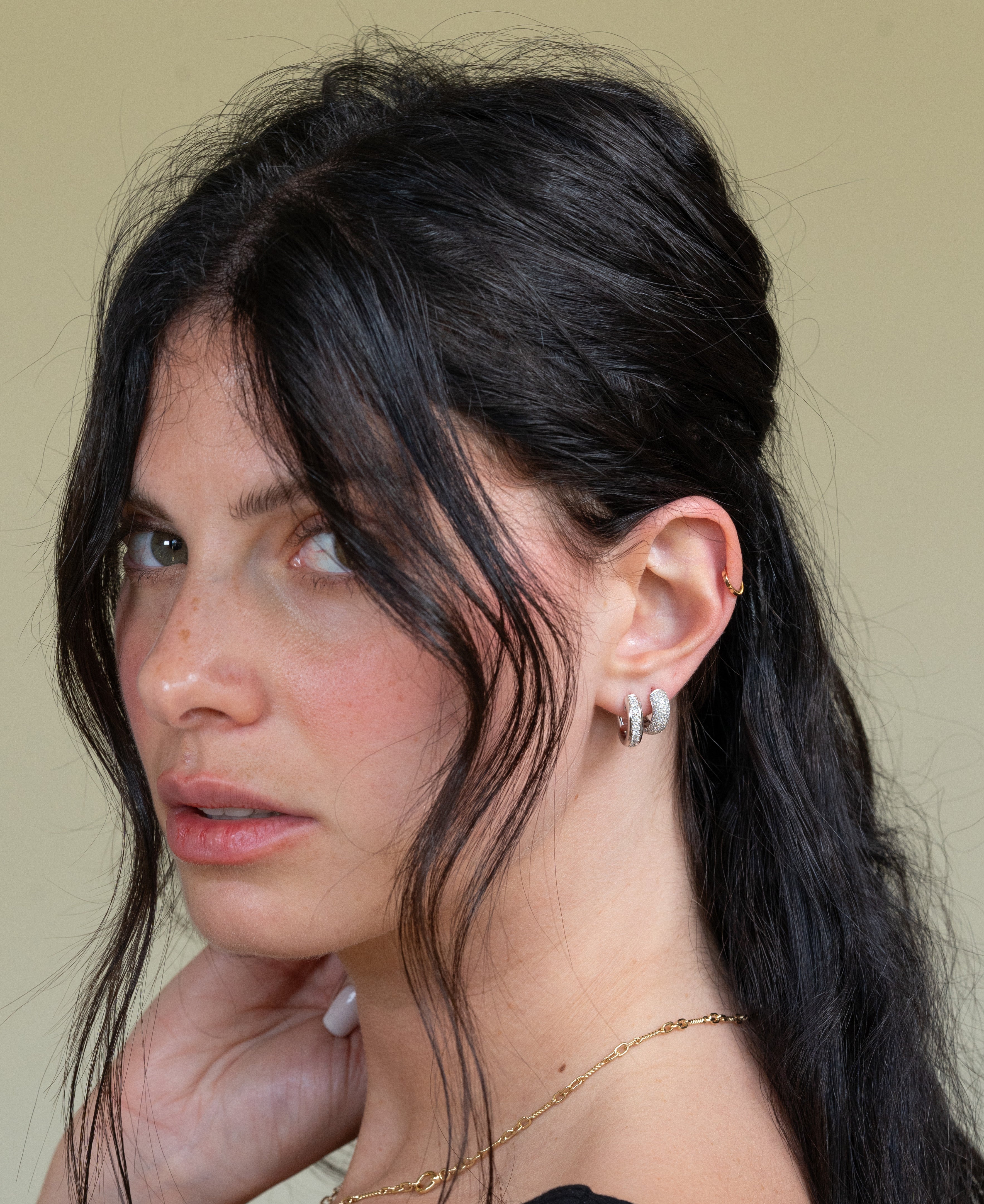 A woman with long black hair looks over her shoulder, highlighting the Pave Diamond Tube Hoops. Her half-up hairstyle with loose strands elegantly frames her face, while the black top and soft beige background enhance the jewelrys allure.