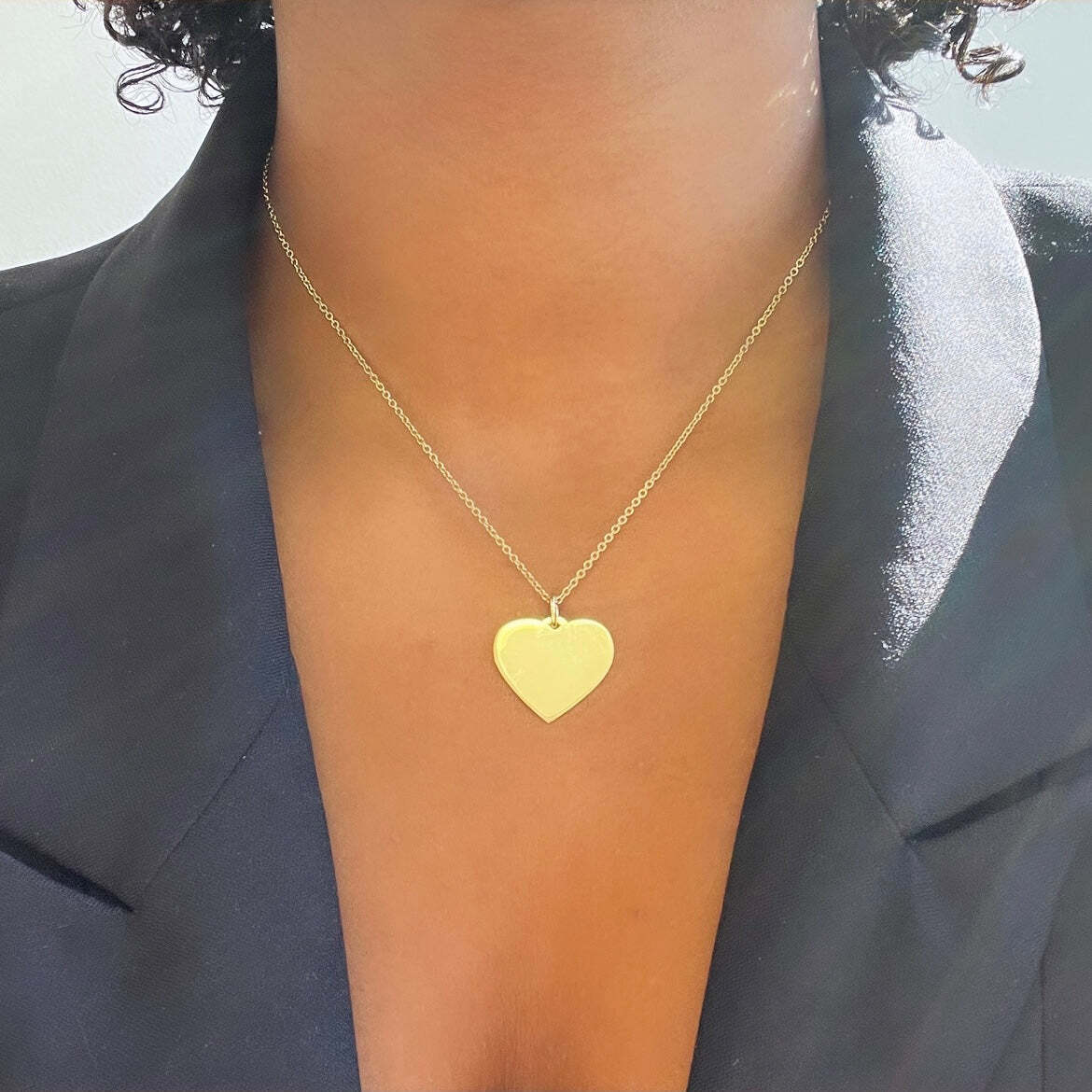 A person wearing the Large Heart Engravable Pendant Necklace, featuring a gold heart-shaped pendant on a thin chain, dressed in a black blazer. The pendant showcases delicate handwriting jewelry, making it a truly personalized necklace and meaningful keepsake.