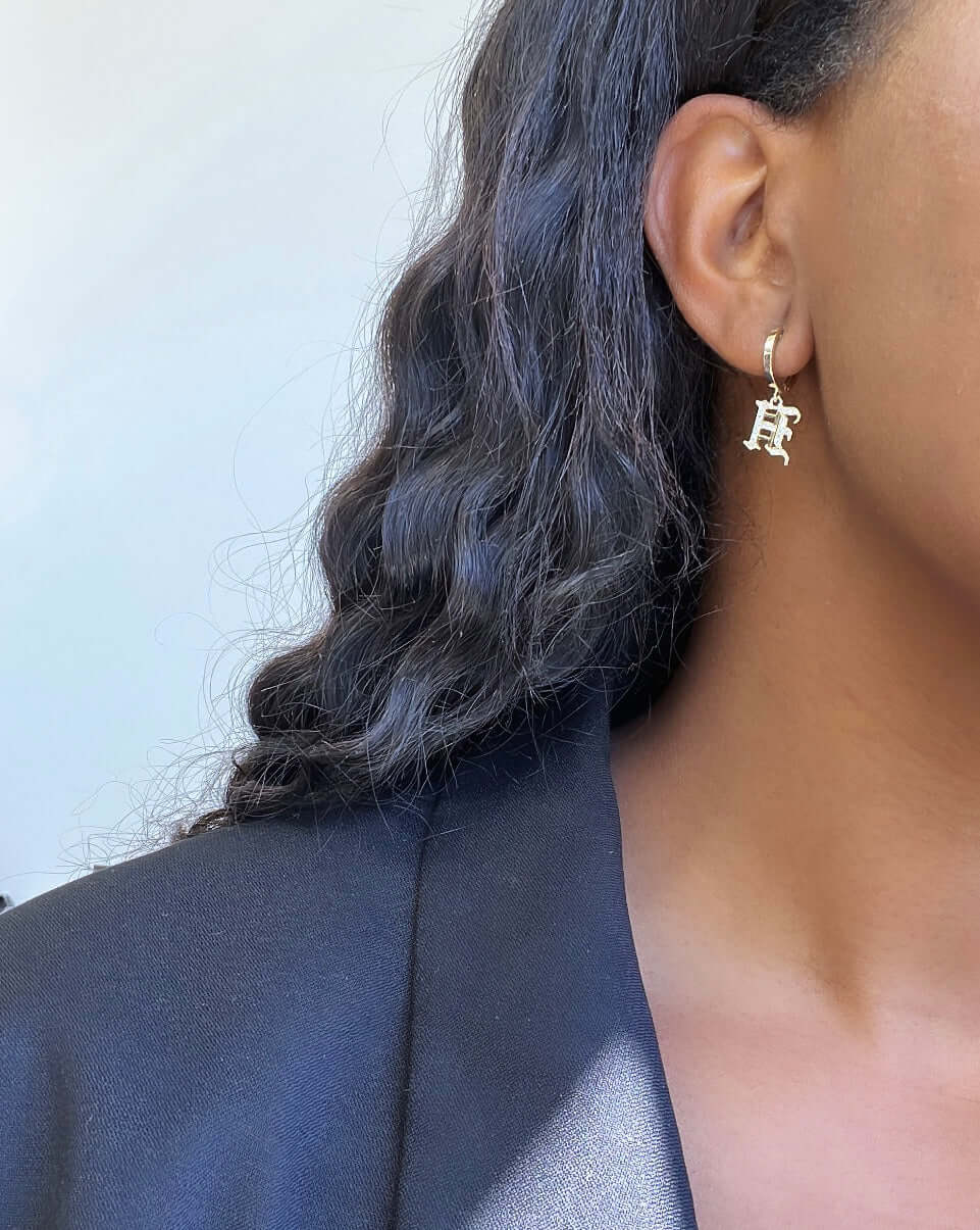 Close-up of a person with long, wavy dark hair wearing a black blazer and the Small Old English Initial Huggies — gold hoop earrings featuring a hanging letter charm. The background is plain and light-colored.