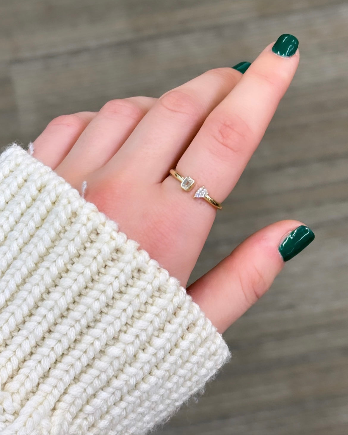 A hand with green nails wearing the Emerald & Pear Toi Et Moi ring, symbolizing love with two shimmering gemstones, rests in a cream knit sweater against a wooden background. Preorder now to add this timeless piece to your collection.