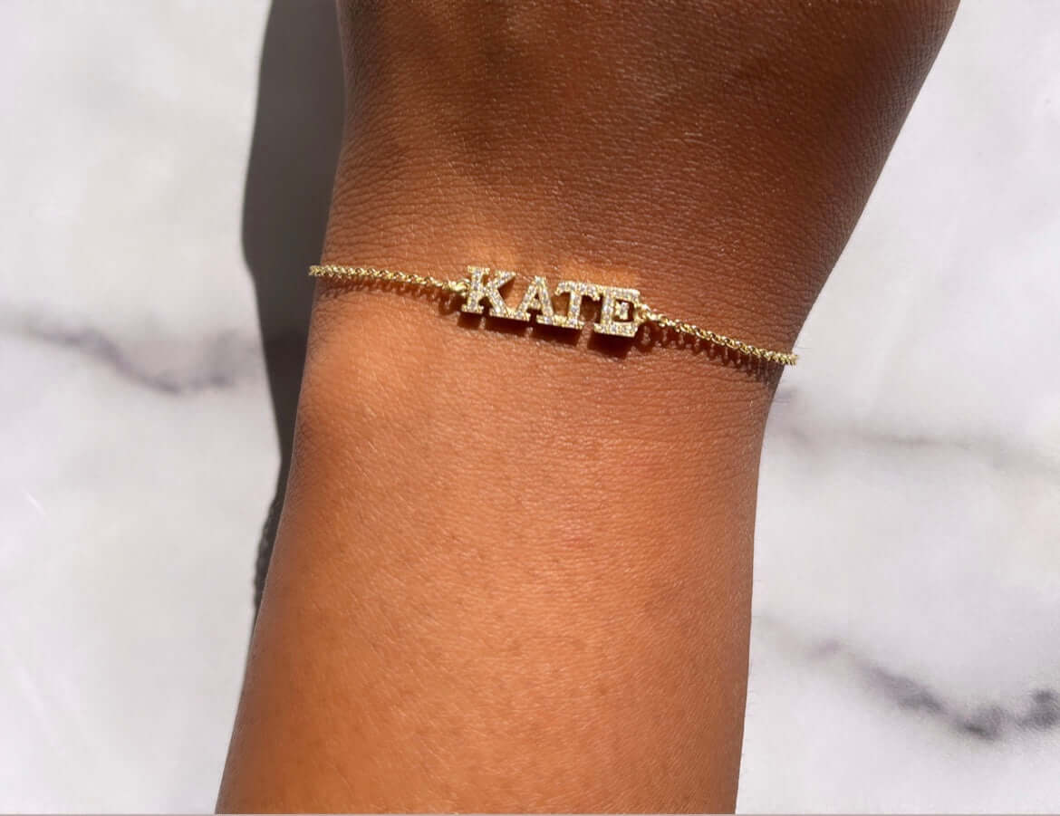 A "Test - Diamond Block Name Bracelet," featuring the name KATE spelled out in diamond-accented block letters, is worn on a person's wrist against a light marble background.