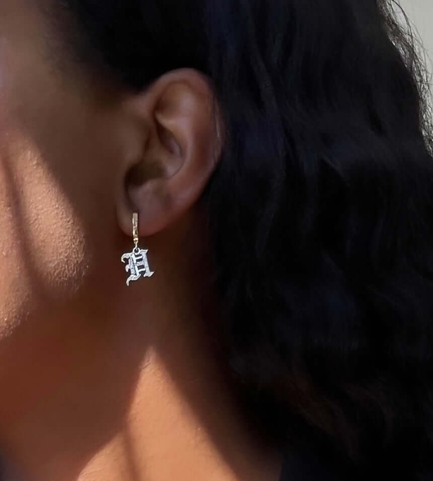 A close-up of a person's ear wearing the "Small Old English Initial Huggies" gold earring. The person has long, dark, wavy hair.