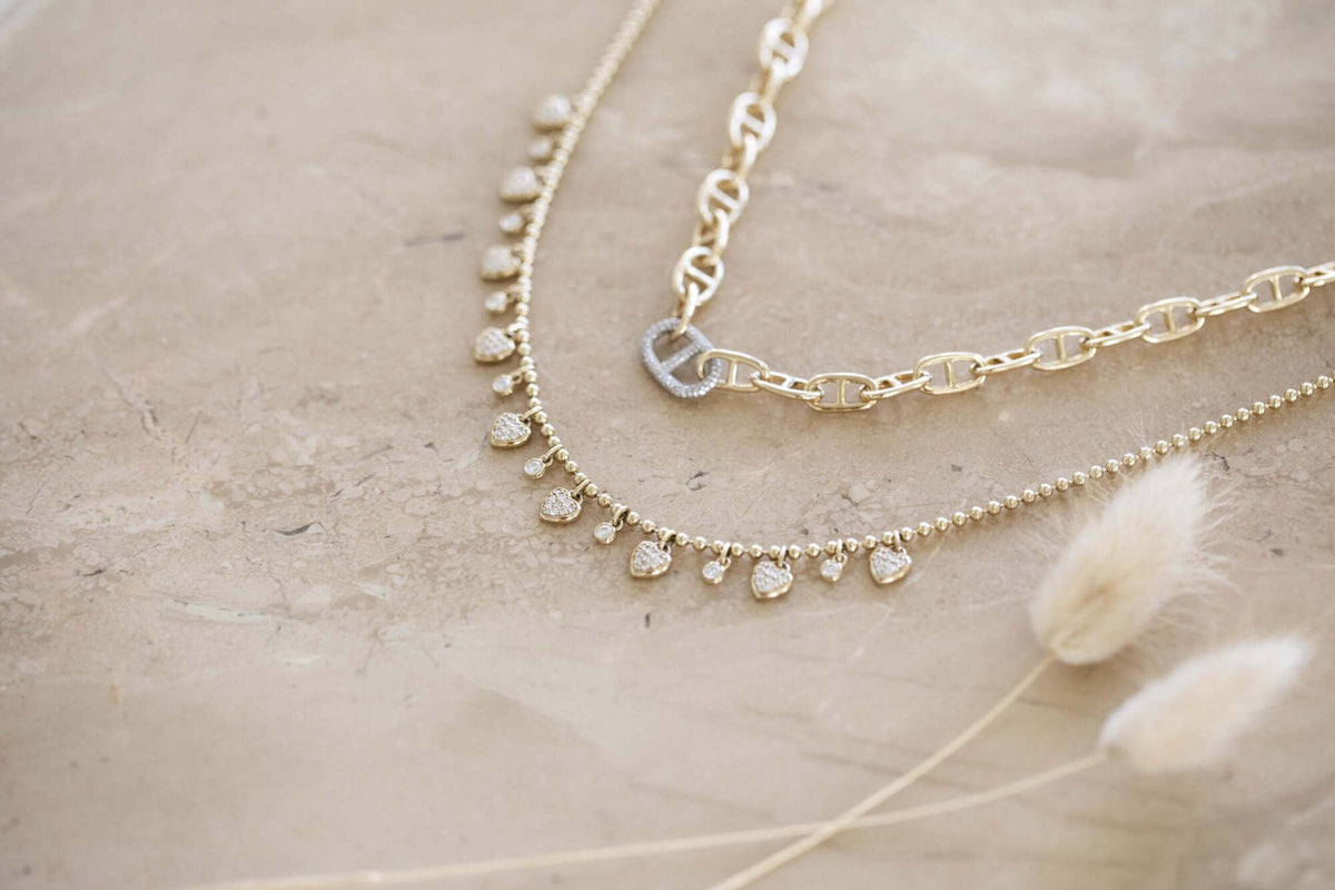 The Diamond Dangle Heart Necklace from Janet Heller Fine Jewelry rests on a beige marble surface, its delicate design featuring dainty diamond-like heart charms. Cream-colored dried grasses nearby underscore the refined elegance of this exquisite fine jewelry collection.