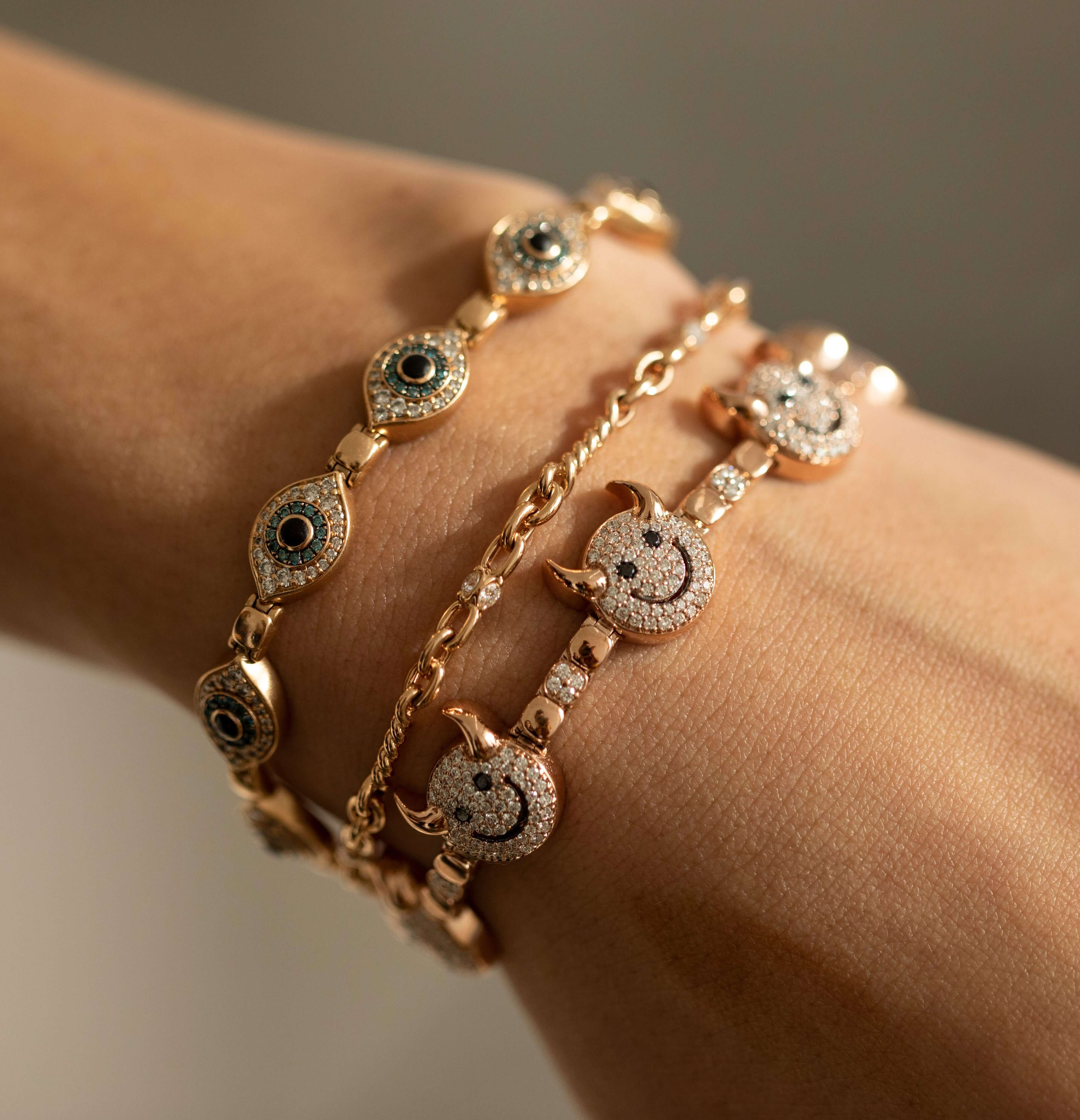 A close-up of a wrist wearing three gold Diamond Belle Bracelets from Janet Heller Fine Jewelry: one adorned with eye-shaped charms, one featuring a simple chain design, and another embellished with crescent moon and star charms, all set with small white and dark stones.