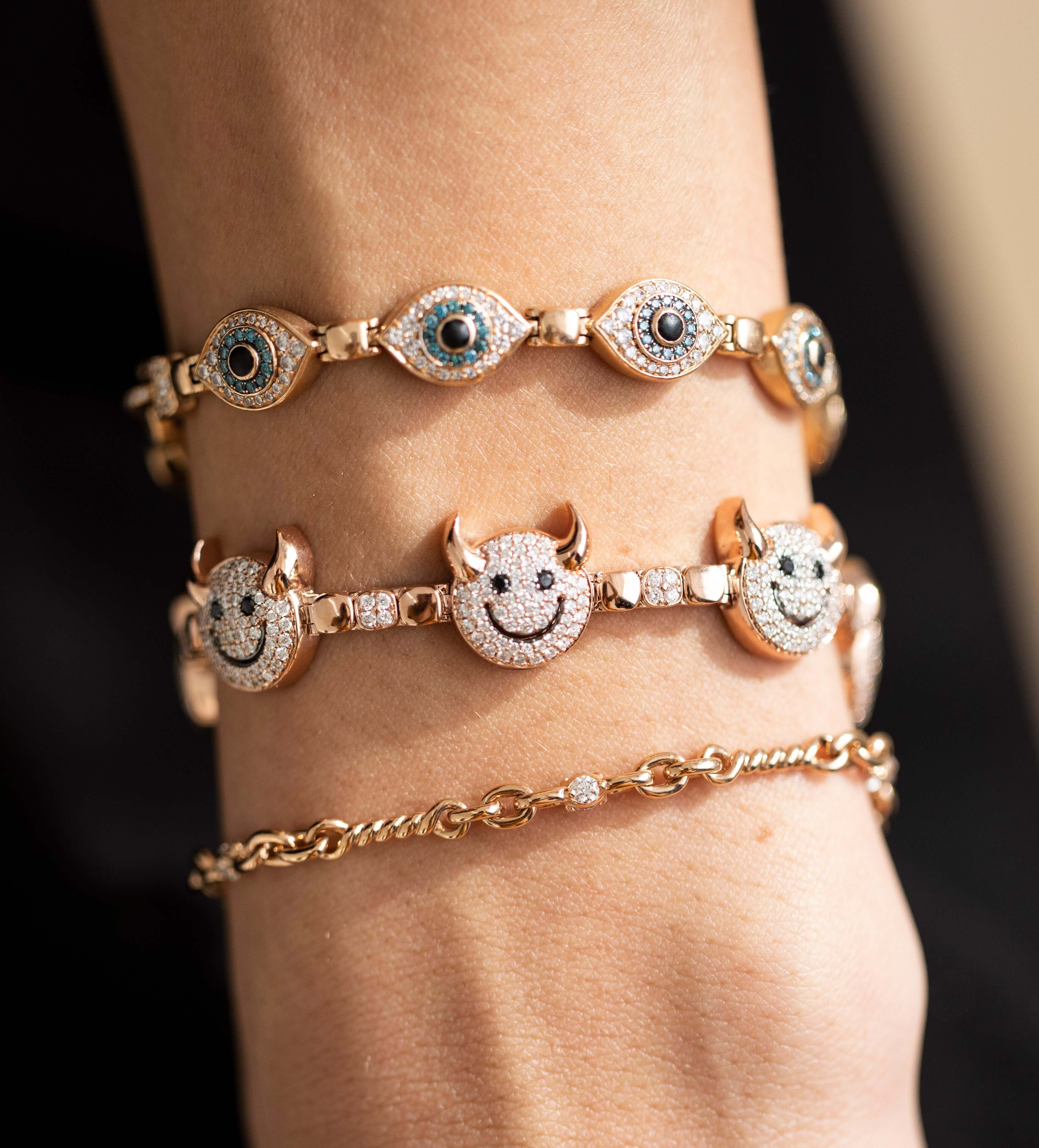 A close-up of a wrist adorned with four gold bracelets from Janet Heller Fine Jewelry, including the Diamond Belle Bracelet featuring a single sparkling gem, alongside designs with evil eye charms, smiling devil faces, and a sleek simple chain.