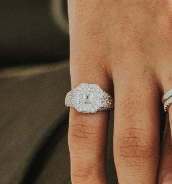 The Diamond Acacia Pinky Ring from Janet Heller Fine Jewelry is crafted in 18KT white gold.  The ring features a geometric design with an emerald-cut center diamond surrounded by smaller pave diamonds in a layered hexagonal setting.