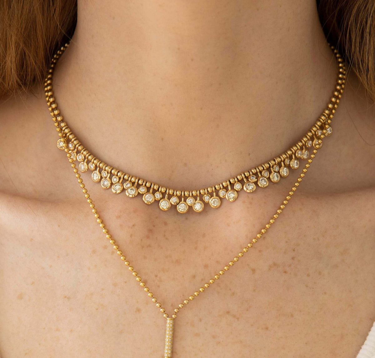 A close-up of a woman's neck highlights two exquisite pieces from Janet Heller Fine Jewelry; one is a fine jewelry choker adorned with small round pendants, while the other is the Diamond Dangle Toilet Chain Necklace, featuring a sleek vertical bar pendant on a longer custom jewelry chain.