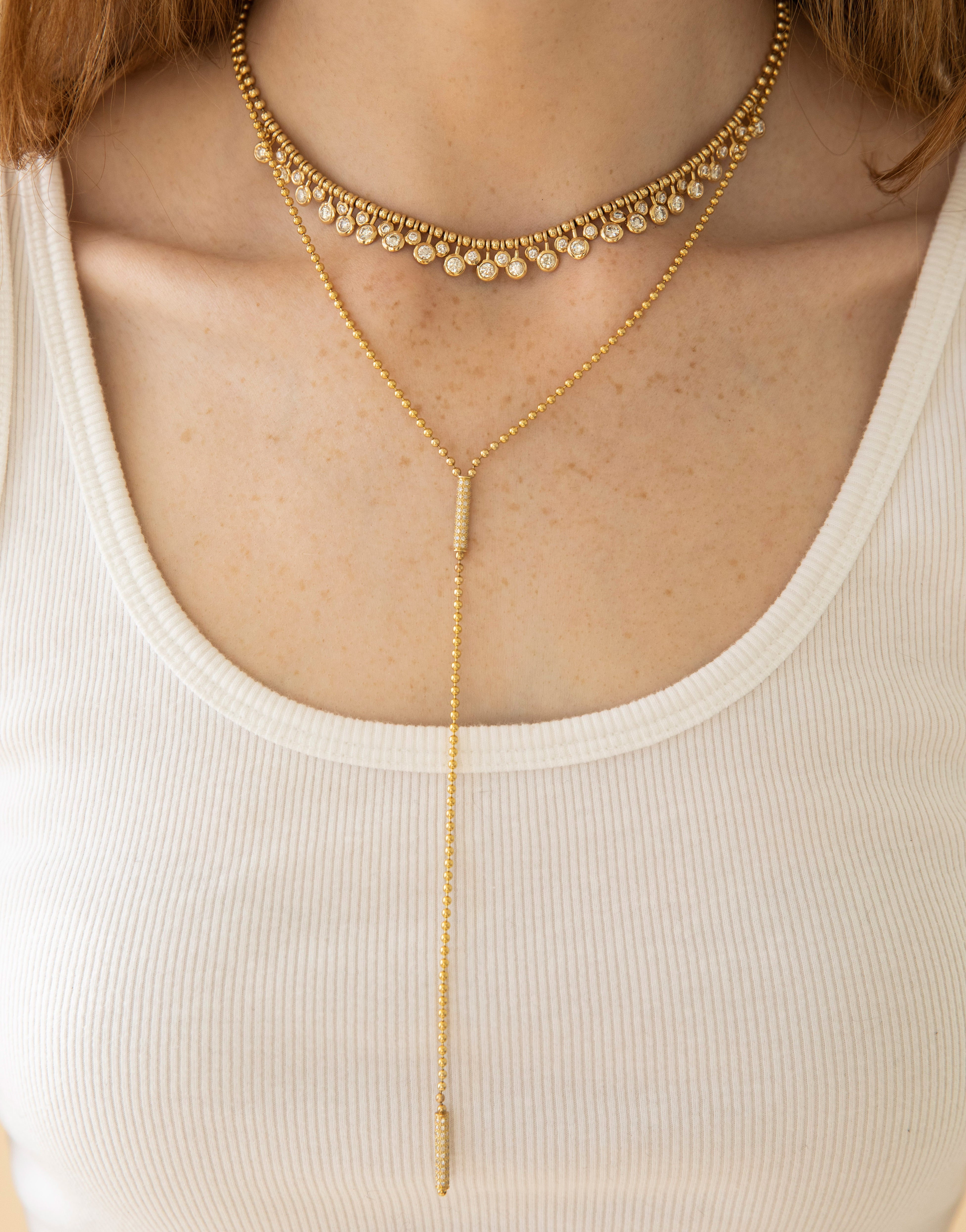 A person in a white ribbed tank top elegantly showcases Janet Heller Fine Jewelry with two layered gold necklaces: one has small circular pendants, and the other is the Toliet Chain Diamond Barrels Lariat featuring a long, thin chain with a vertical drop.