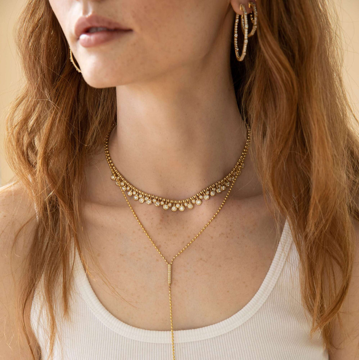 A woman with wavy auburn hair wears a white tank top and layered gold necklaces—one featuring the Diamond Dangle Toilet Chain Necklace from Janet Heller Fine Jewelry, paired with another shorter charm necklace and gold hoop earrings. Her lips and neck are visible, but her eyes are not shown, drawing attention to the elegance of this fine jewelry selection.