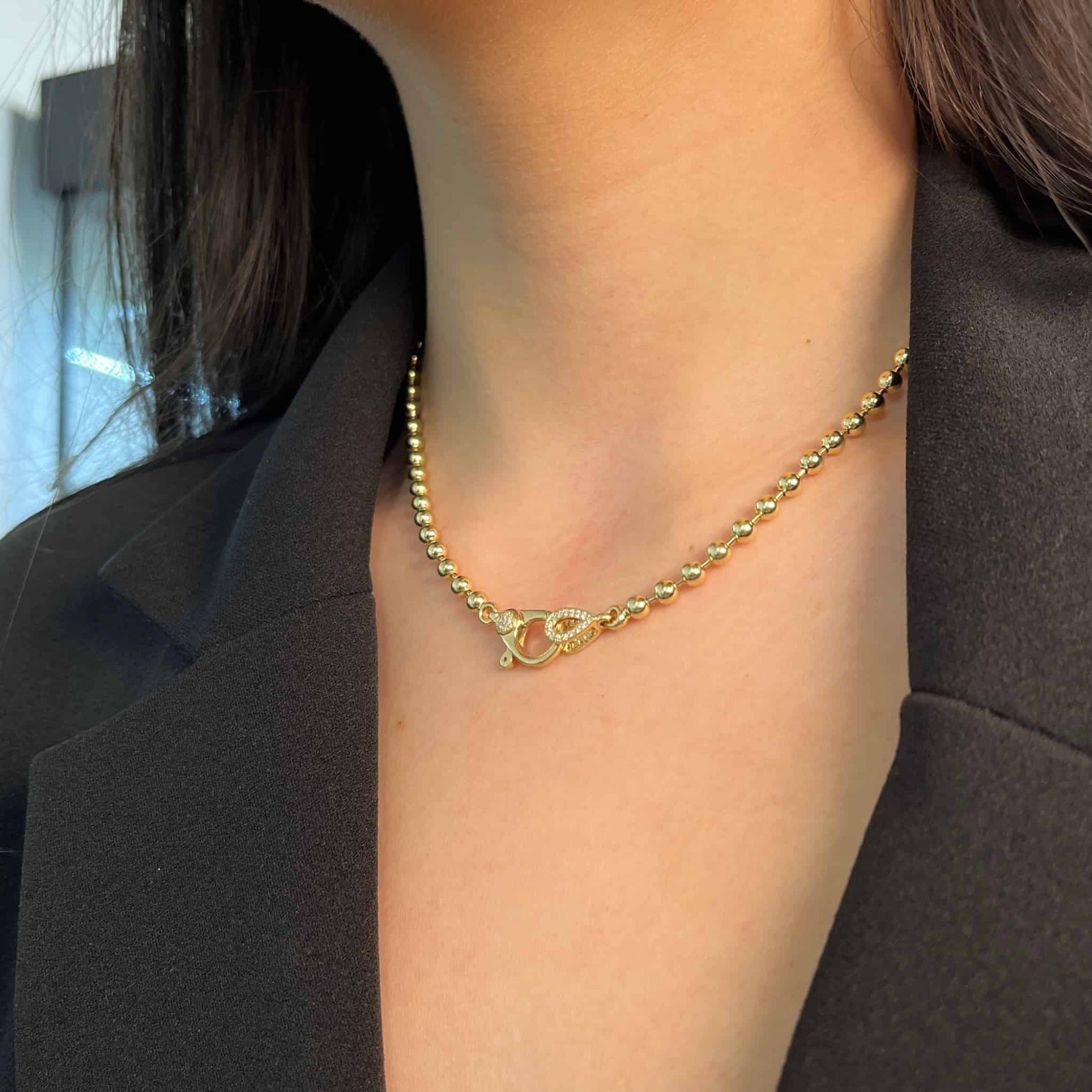 A woman wearing the 4mm Paris Chain with a heart-shaped clasp, dressed in a black blazer, showcases the timeless elegance of Janet Heller Fine Jewelry. The delicate gold beading and charming design of the necklace add sophistication to her ensemble.