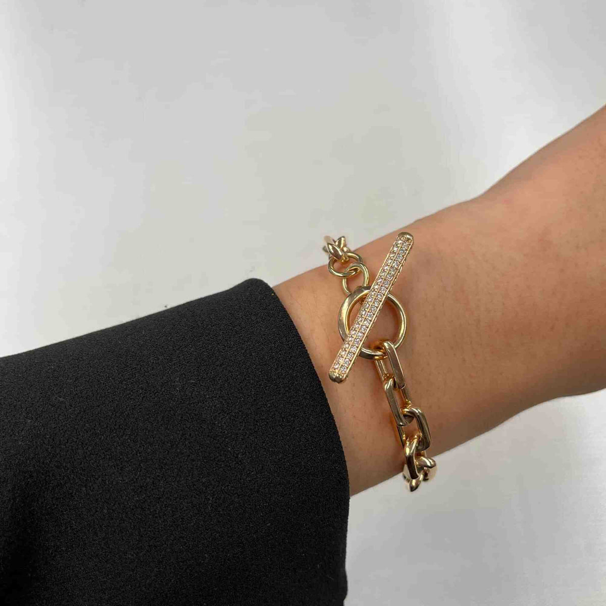 A persons wrist elegantly displays a Full Square Diamond Toggle Link Bracelet, boasting a gold chain and toggle clasp adorned with crystals by Janet Heller Fine Jewelry. Dressed in a black sleeve, they pose against a plain light grey surface.