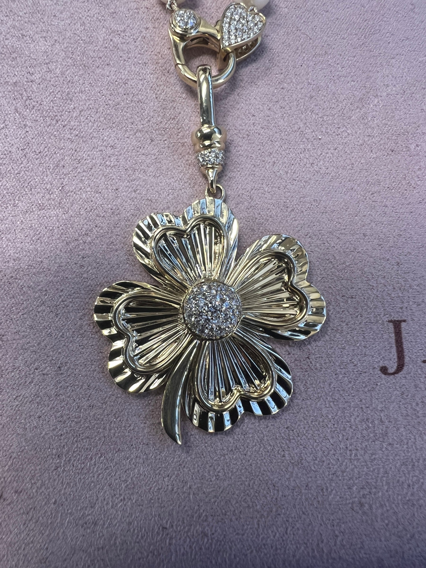 The Jumbo Diamond Clover Charm is a gold, flower-shaped pendant with a textured surface and a cluster of small diamonds at its center, shown on a soft, light purple background.