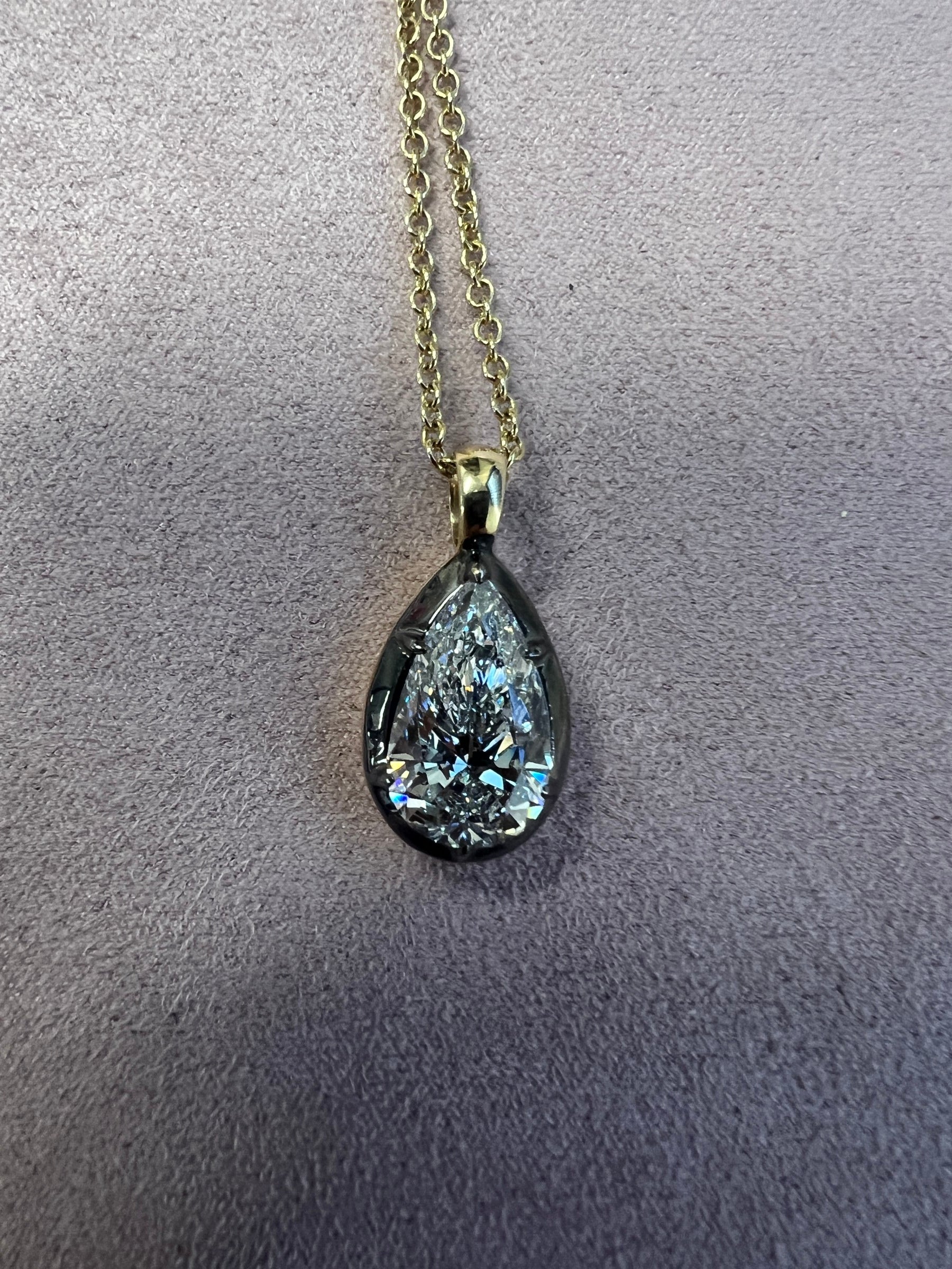 The Lab Grown Pear Diamond Black Rhodium Charm features a sparkling pear-shaped lab grown diamond pendant on a gold chain, displayed on a light gray textured surface.