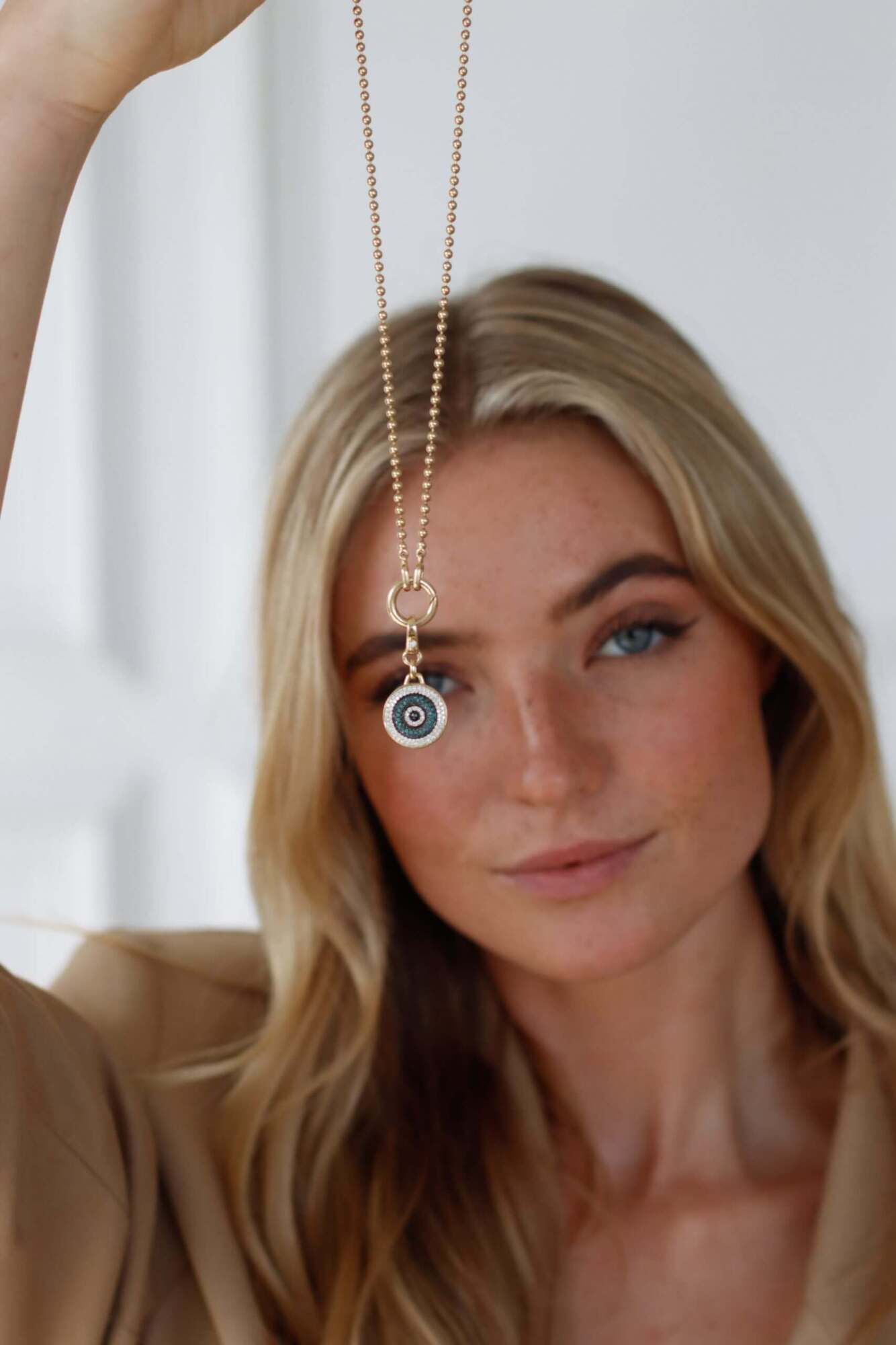 A woman with long blonde hair holds up the Amsterdam Chain by Janet Heller Fine Jewelry, featuring a striking blue evil eye pendant and delicate jewelry charms, focusing on the intricate details while her face appears slightly out of focus in the background.