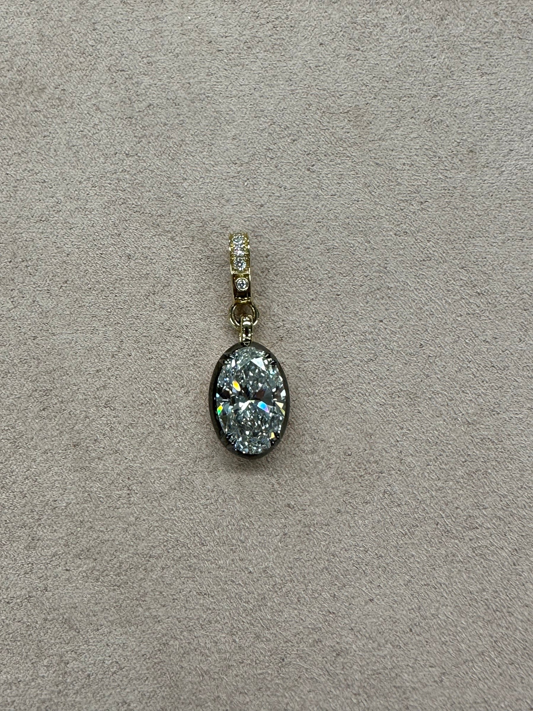 The Oval Lab Diamond Charm with Black Rhodium features a sparkling diamond in a gold bezel and smaller diamonds on the bail, showcased on a beige textured background. From Janet Heller Fine Jewelry.