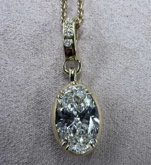 The Oval Lab Grown Diamond Charm features an oval diamond pendant in a gold frame with a round lab grown diamond accent above it, all set against a lightly textured fabric background.