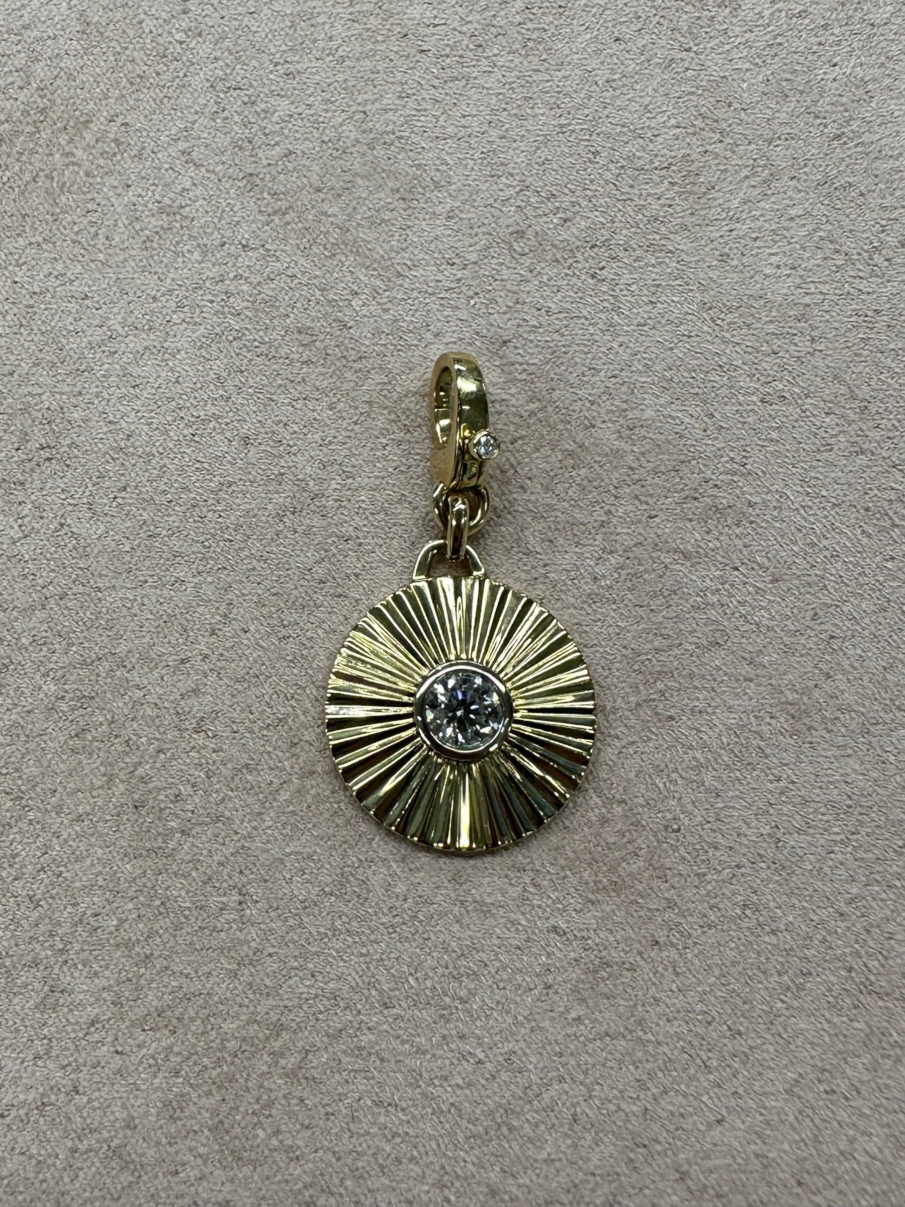 The Ribbed Diamond Cut Wheel Charm features radiating lines and a central clear gemstone, crafted in 14KT gold and finished with a gold lobster clasp. Displayed on a beige textured surface, this charm is an elegant fine jewelry piece.